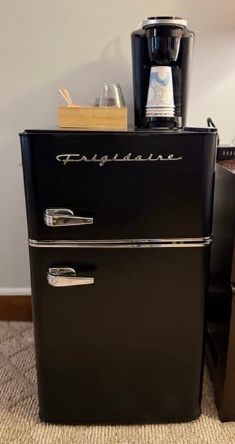 A close-up of a black retro-style Frigidaire mini-fridge with silver handles and trim. A black Keurig coffee maker sits on top of the fridge, alongside a wooden organizer holding coffee accessories.