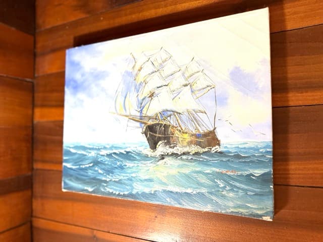 A close-up of a framed painting of a traditional sailing ship with multiple white sails navigating through choppy blue ocean waves under a soft, cloudy sky. The painting is mounted on a warm, natural wood-paneled wall.