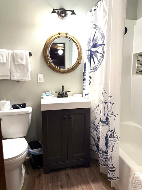 A brightly lit bathroom featuring a dark wood vanity with a white countertop and a brass-colored porthole-style mirror mounted on a light gray wall. To the right, a white shower curtain with a navy blue nautical compass and ship illustration is visible, alongside a white bathtub and toilet.