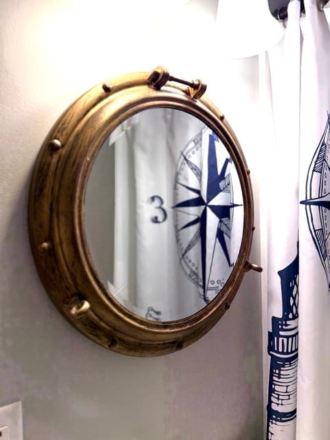 A brightly lit bathroom featuring a dark wood vanity with a white countertop and a brass porthole-style mirror mounted on a light gray wall. To the right, a white shower curtain with a navy blue nautical compass and ship illustration is partially visible.