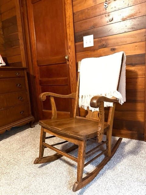 A classic wooden rocking chair with a white fringed throw blanket draped over the back, situated on a carpeted floor against warm, natural wood-paneled walls. A dark wood dresser is partially visible in the background.