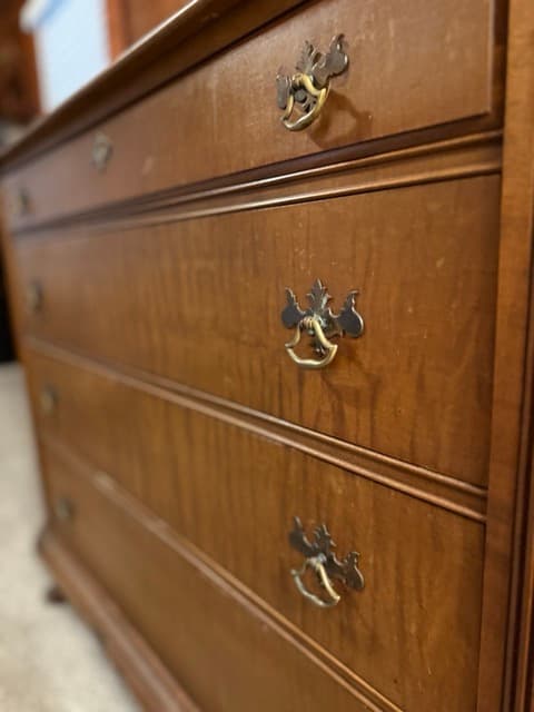 A close-up, angled view of a vintage-style wooden dresser with a warm finish. The dresser features several drawers adorned with decorative, ornate brass-colored metal pull handles.