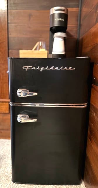 A close-up of a black retro-style Frigidaire mini-fridge with silver handles and trim, set against a warm wood-paneled wall. A black Keurig coffee maker and a wooden accessory organizer sit on top of the unit.