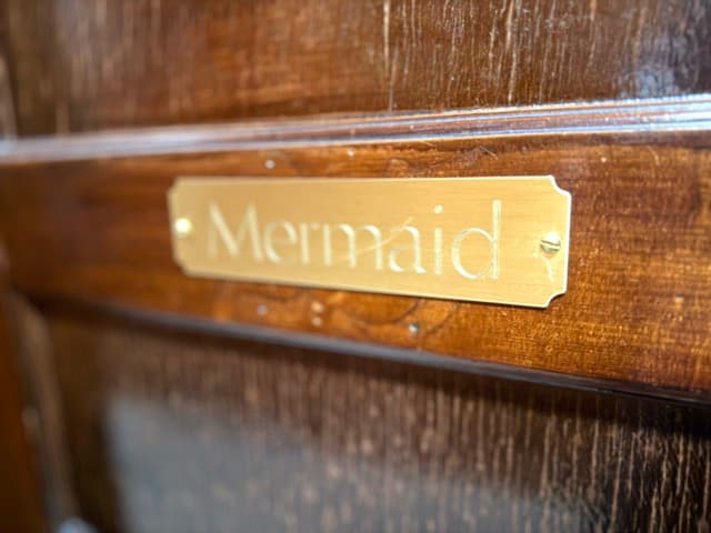 A close-up of a polished brass nameplate engraved with the word "Mermaid" in a serif font, mounted on a dark, grain-textured wooden door.