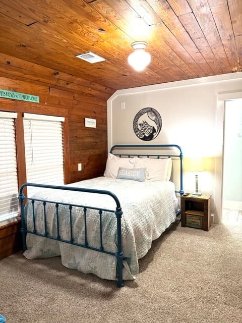 A nautical-themed guest room featuring a blue metal-frame bed with white bedding and a light blue accent pillow. One wall is covered in rich natural wood paneling, while the adjacent wall is a light gray and features a circular black-and-white illustration of a mermaid. The room is warmly lit by a ceiling globe light and a bedside lamp on a small wooden nightstand.
