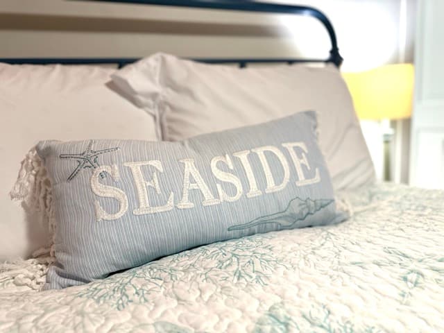 A close-up of a light blue decorative pillow with white tasseled ends resting on a white patterned quilt. The pillow is embroidered with the word "SEASIDE" in white capital letters and features subtle nautical illustrations of a starfish and a seashell. The dark metal frame of a bed is visible in the blurred background.