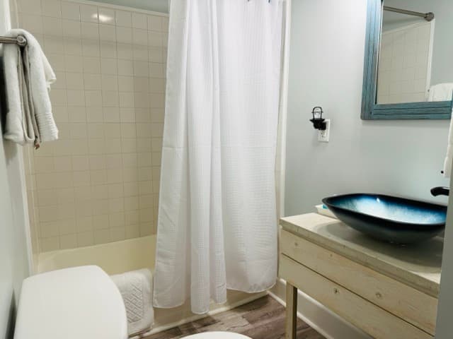 A modern bathroom featuring a light wood vanity with a dark, bowl-style vessel sink and a black faucet. The room includes a white bathtub with a white shower curtain, light gray walls, a blue-framed mirror, and wood-style flooring.