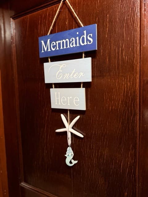 A decorative wooden hanging sign on a dark door. The sign consists of three stacked plaques connected by rope that read "Mermaids," "Enter," and "Here" in white text, accented by a white starfish and a small mermaid figurine dangling at the bottom.