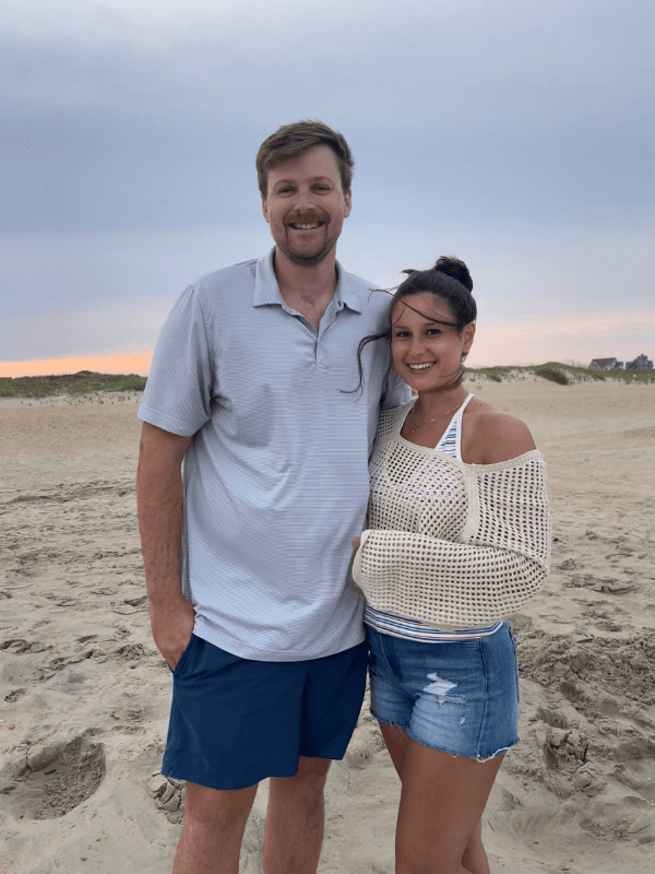 A man and a woman stand smiling together on a beach at sunset.