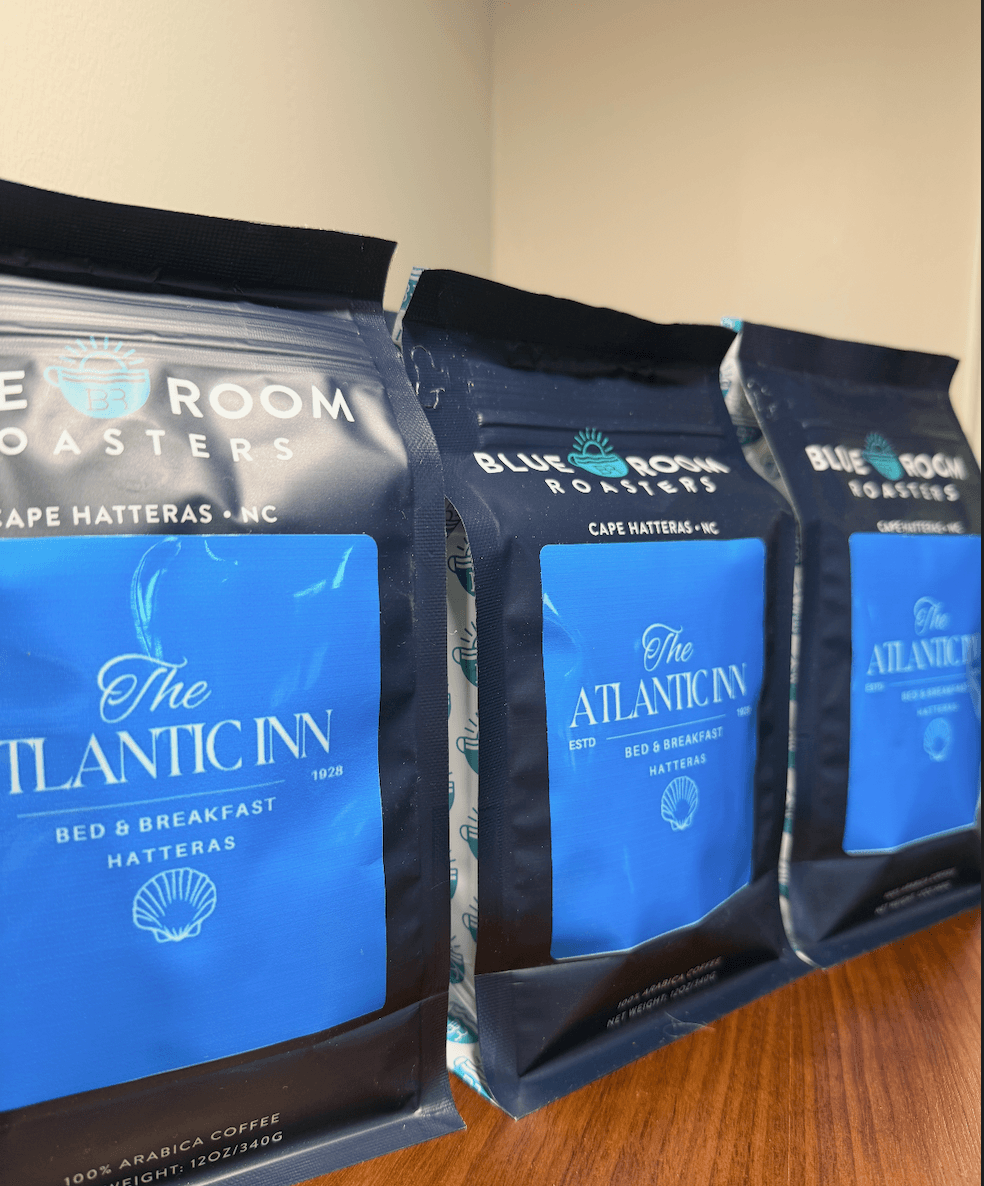 Three bags of Blue Room Roasters coffee featuring "The Atlantic Inn, Hatteras" label, arranged on a wooden surface.