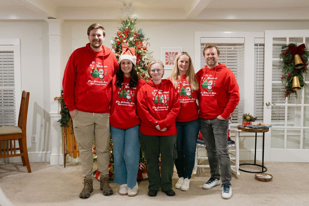 A group of five people stands in front of a Christmas tree, all wearing matching red holiday sweatshirts.