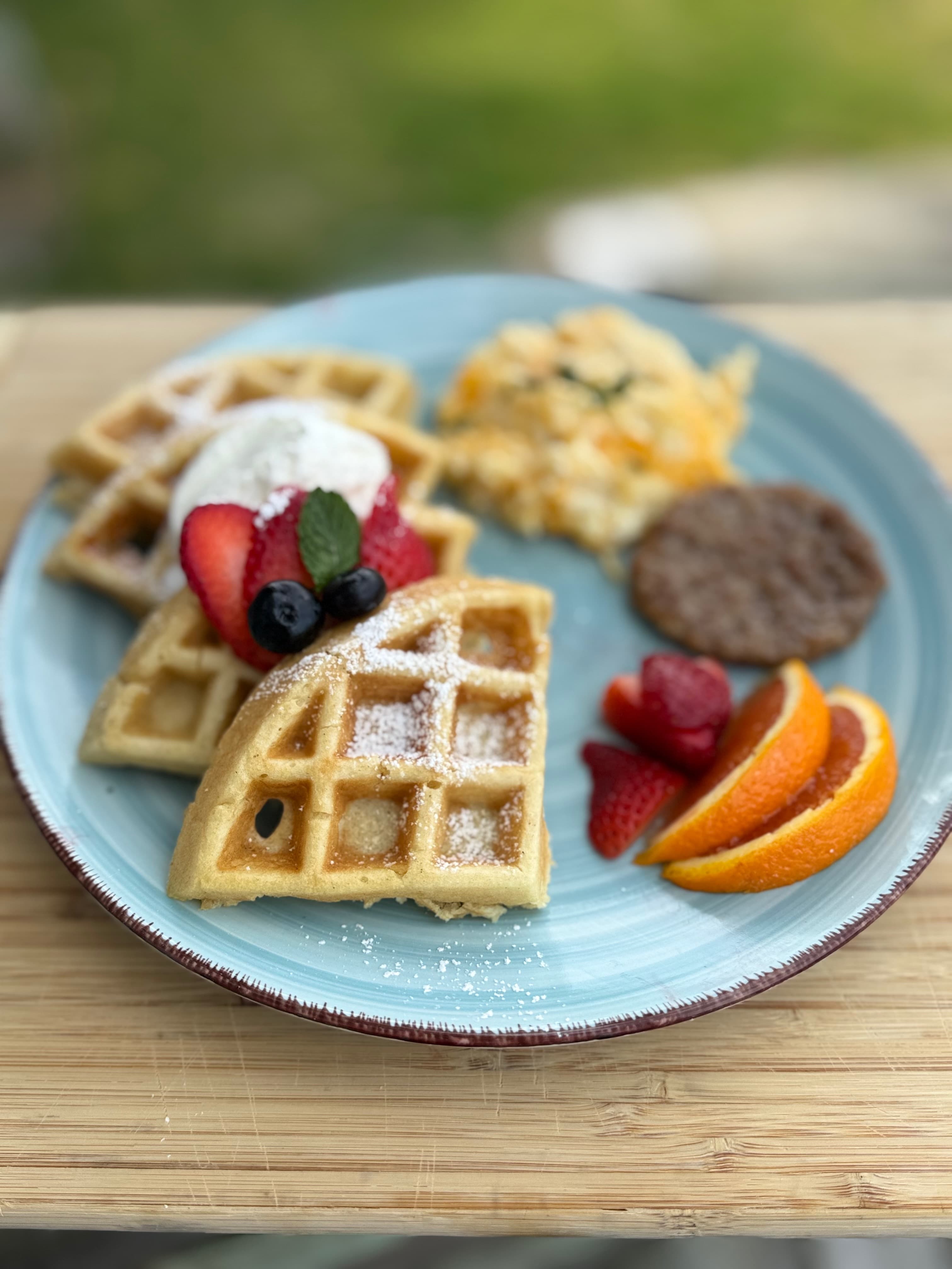 A plate of waffles topped with berries and whipped cream, accompanied by scrambled eggs, sausage, and orange slices.