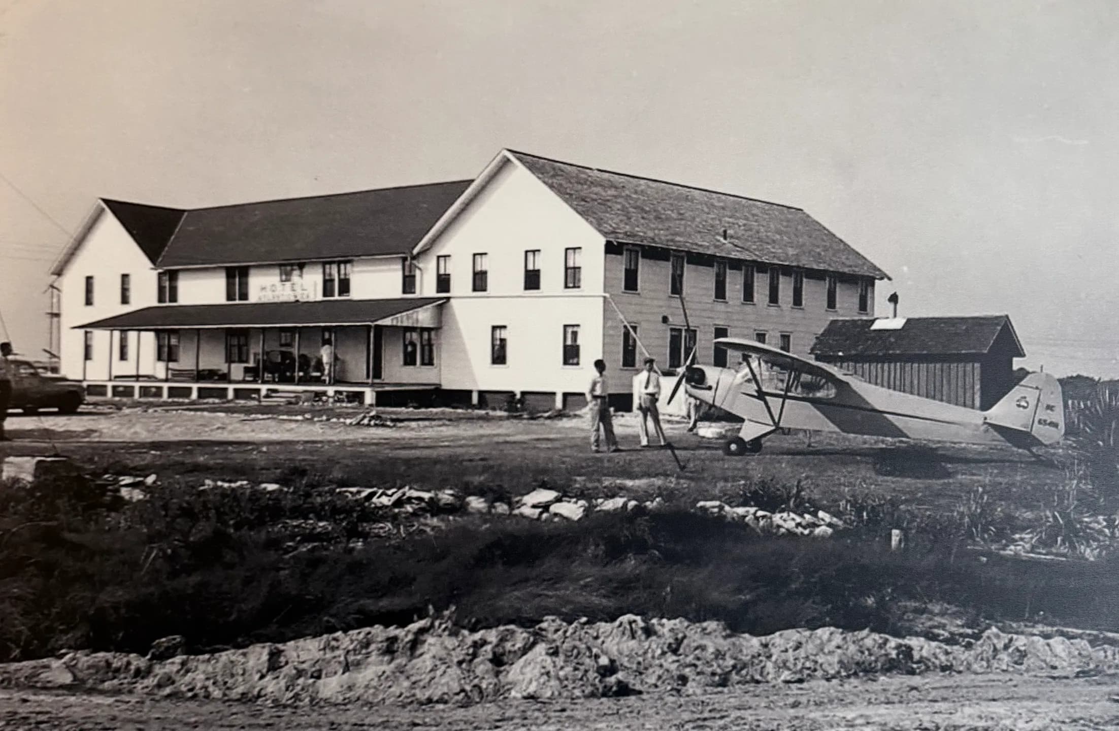 A historic black-and-white photo of a large building with a small airplane and people standing nearby.