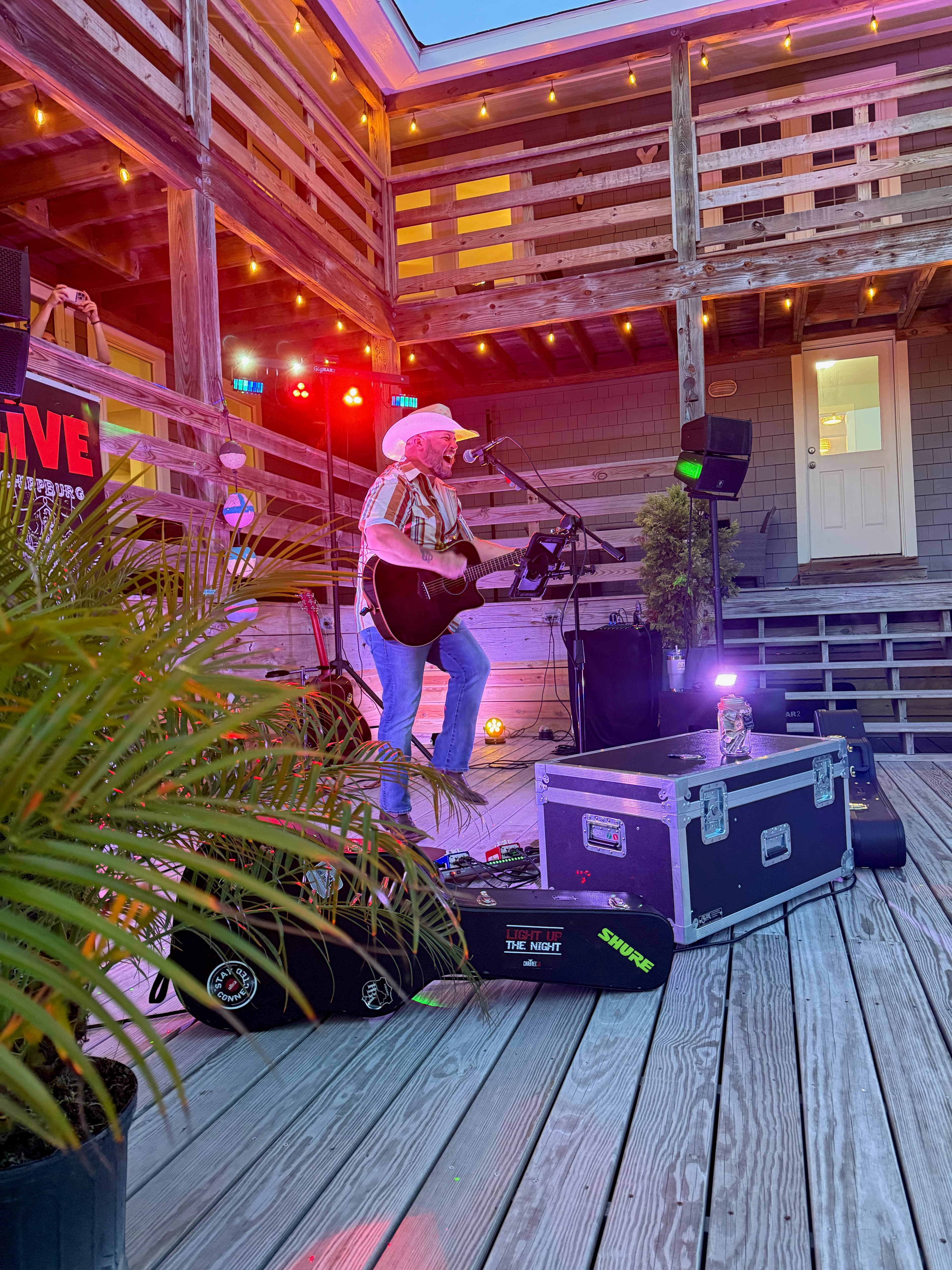 A musician in a cowboy hat performs on an outdoor stage with colorful lights and musical equipment.