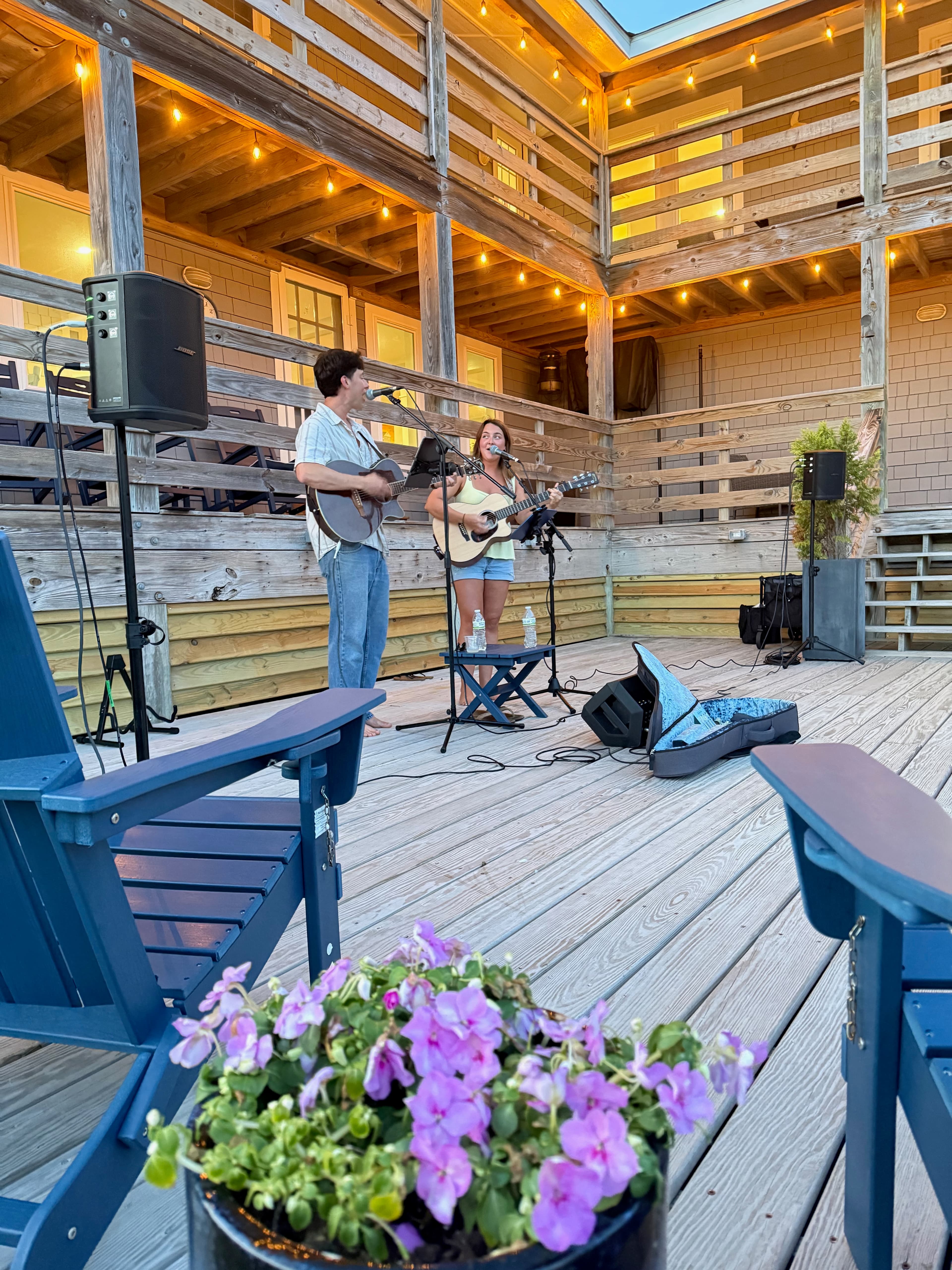 A man and a woman perform live music on a wooden deck, with flowers and chairs in the foreground.