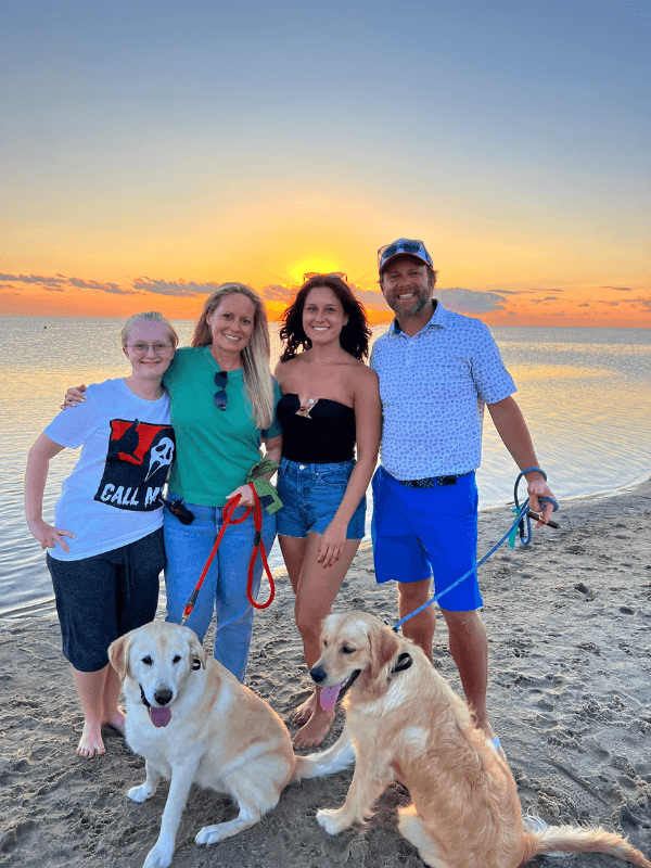 A group of four people with two dogs stands on the beach at sunset.