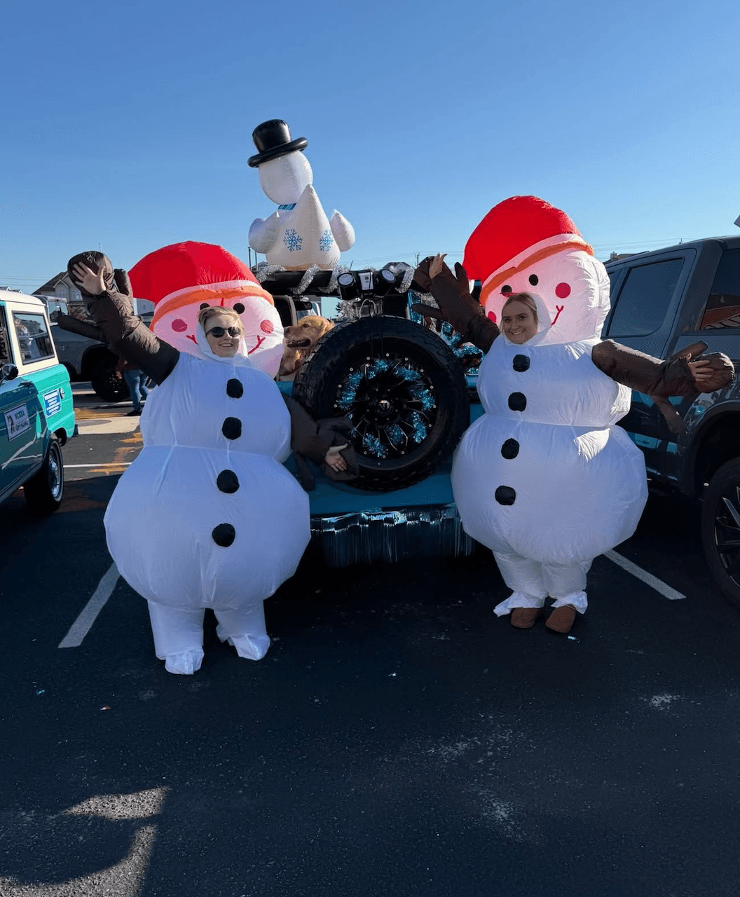 Two women in inflatable snowman costumes pose joyfully in front of a decorated vehicle with a snowman prop.