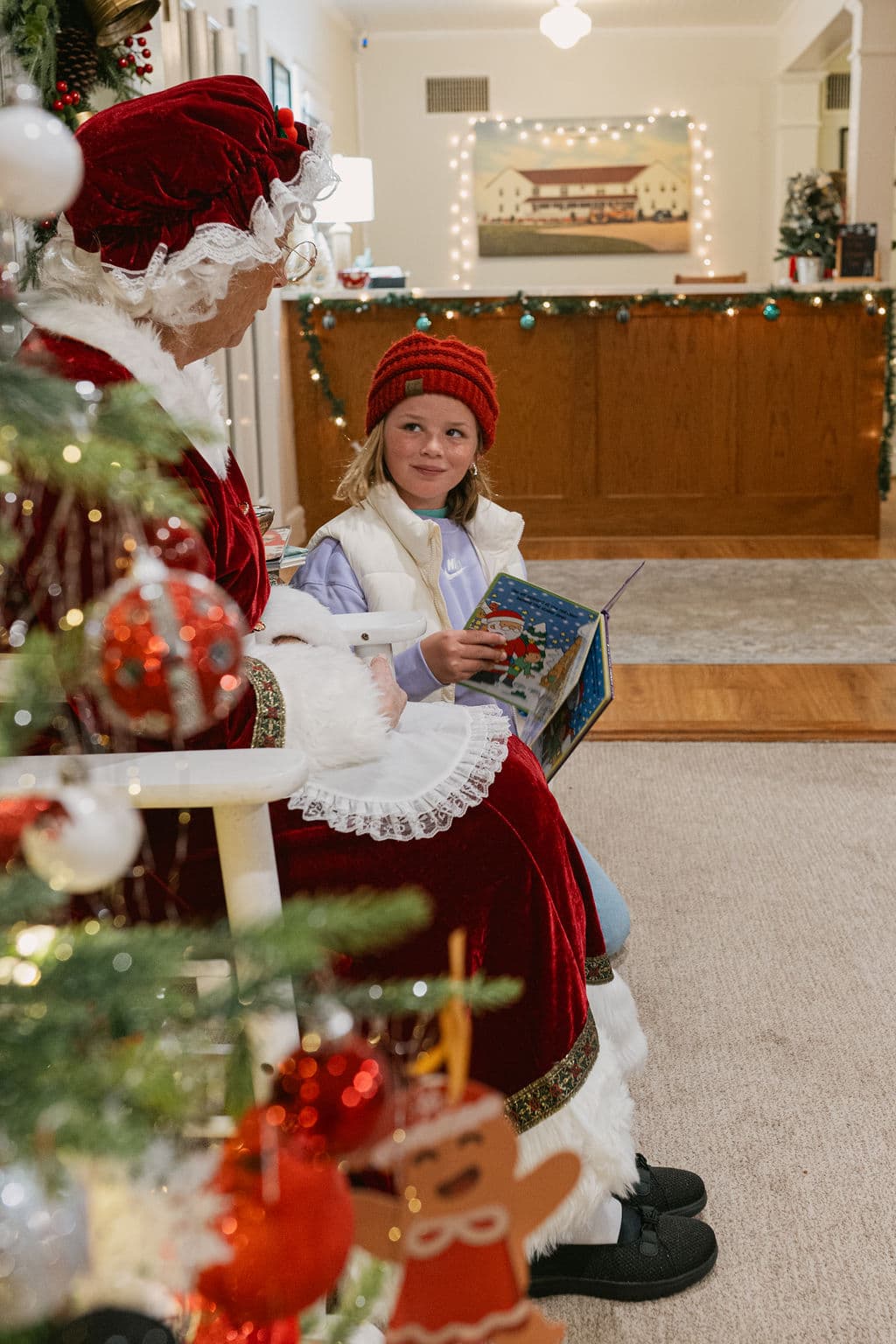 A smiling child in a red hat reads a book while sitting next to a person dressed as Mrs. Claus in a festive setting.