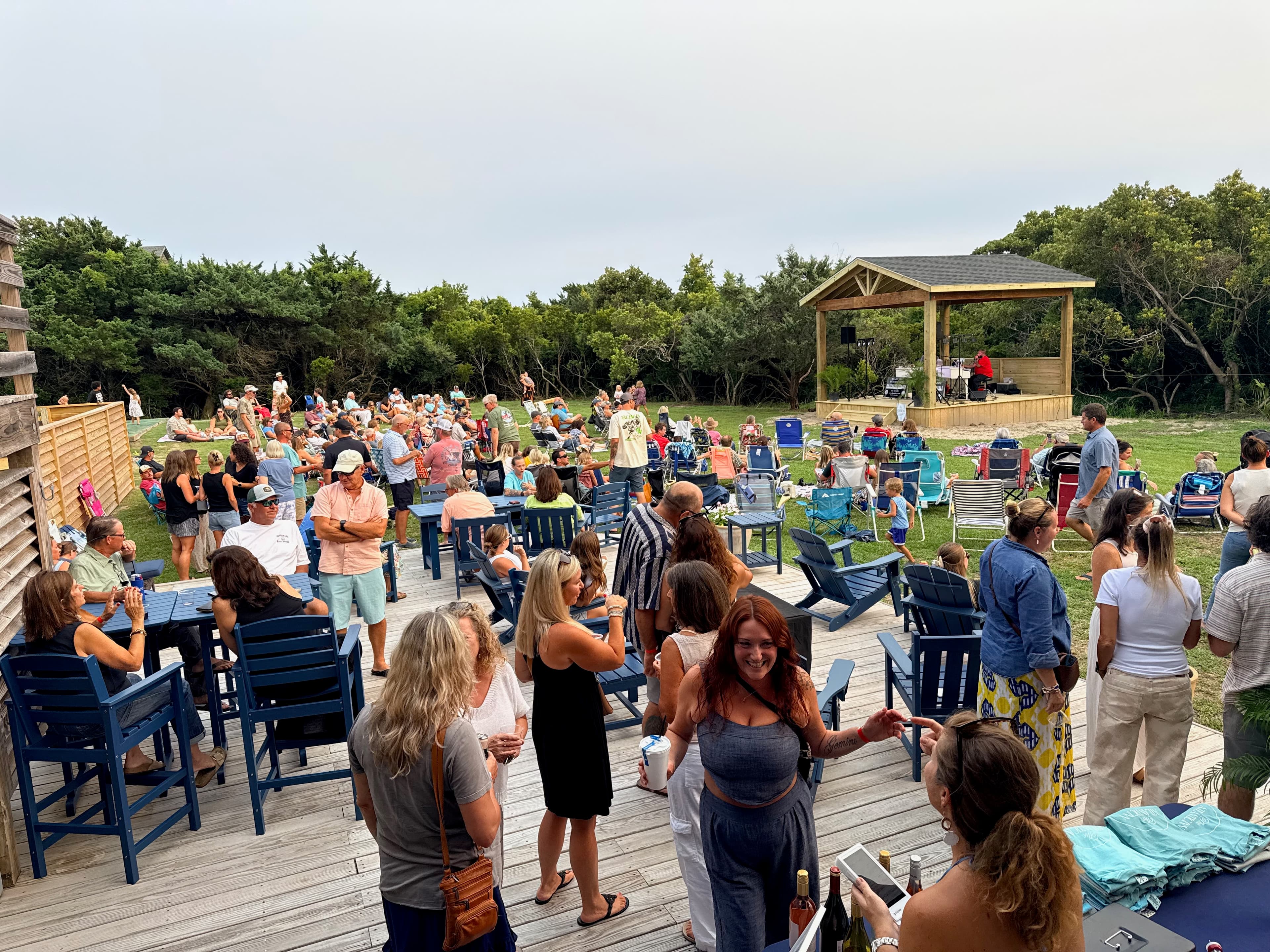 A lively outdoor gathering with numerous people socializing in a grassy area surrounded by trees.