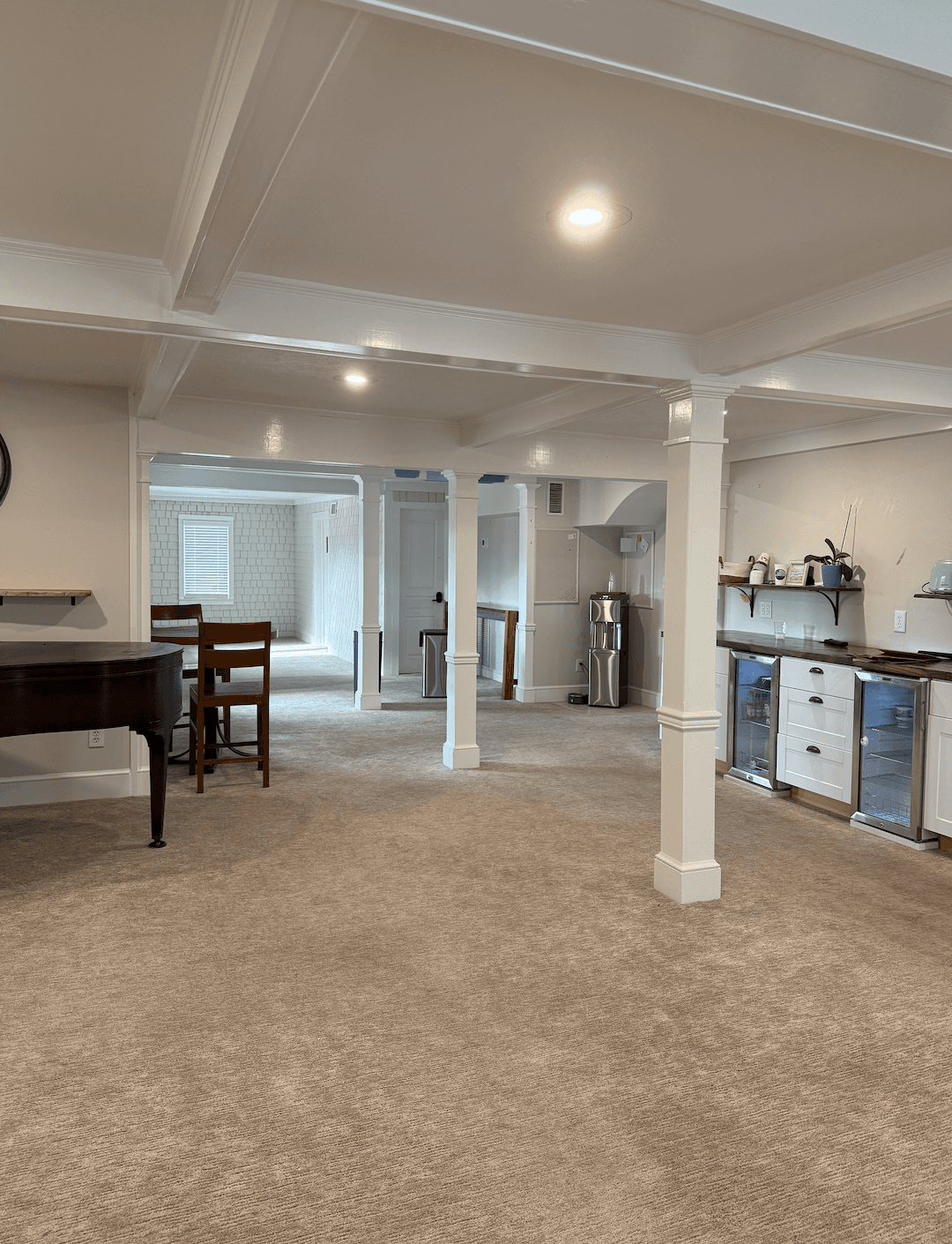 A spacious, carpeted basement featuring white pillars, a piano, and a kitchenette area.