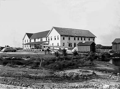 A historic black and white photograph of a large building with an attached aircraft.