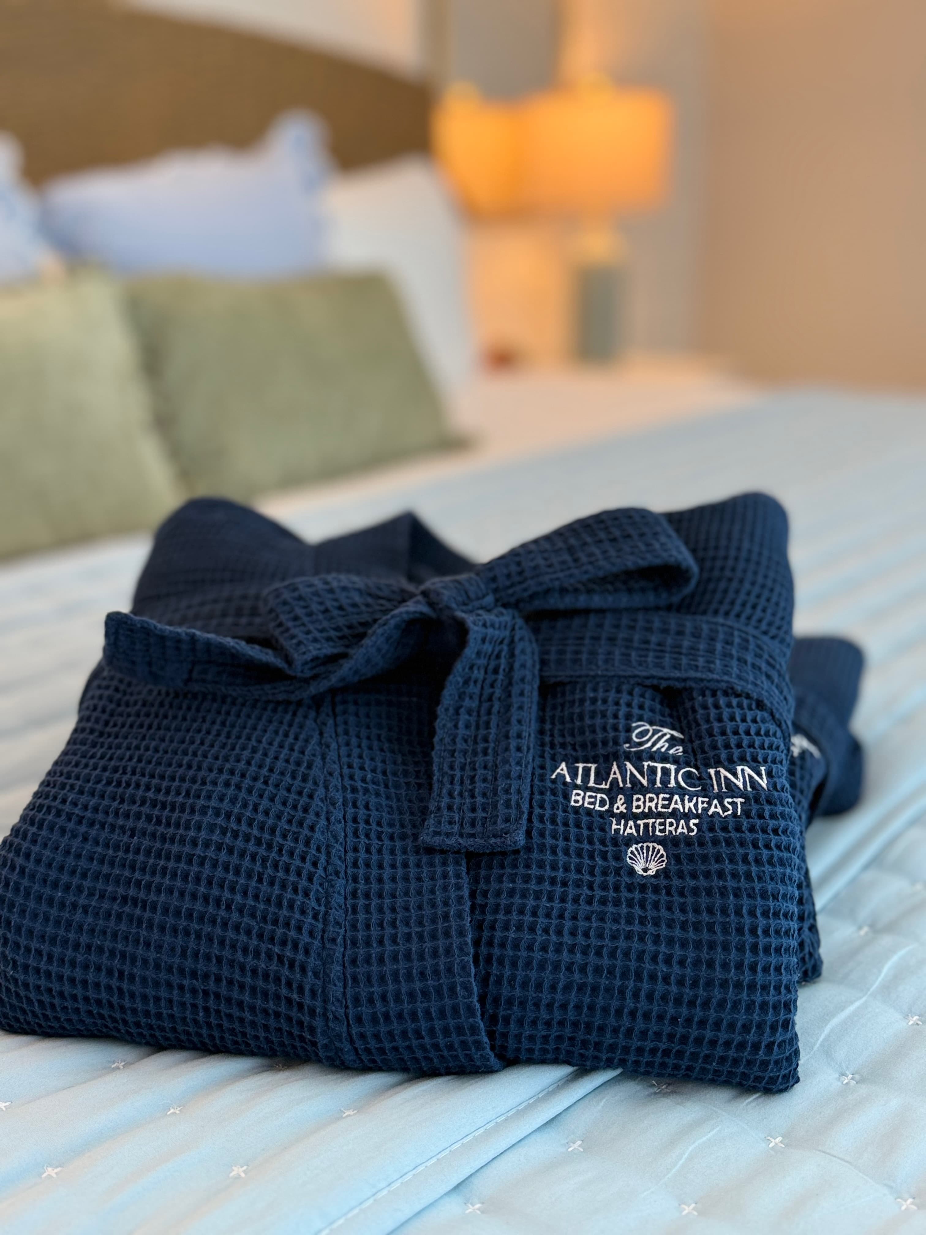 A close-up of two navy blue waffle-weave bathrobes embroidered with "Atlantic" in black script, neatly folded on a bed with blue and white striped bedding.