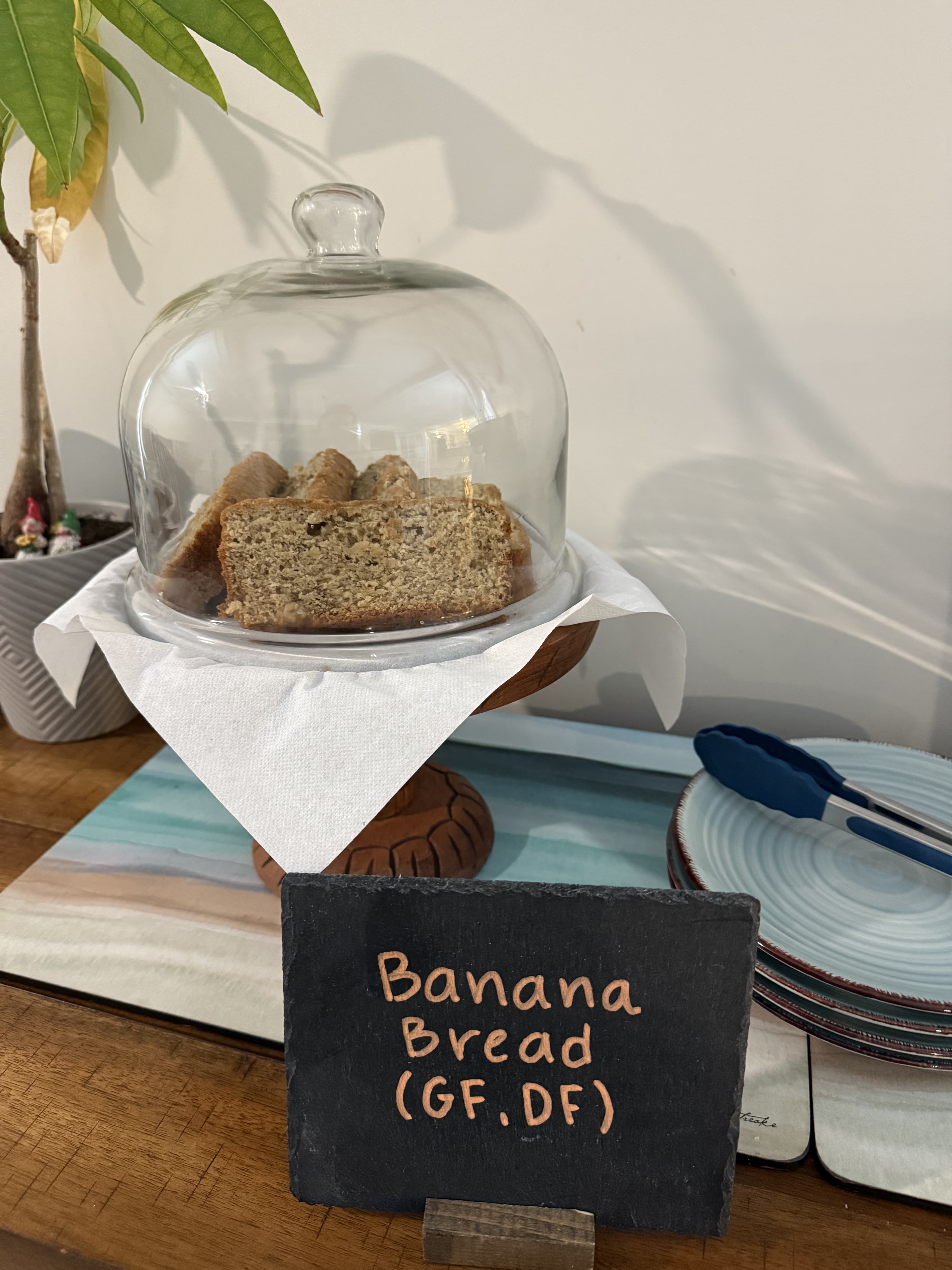 A glass cloche covers slices of gluten-free, dairy-free banana bread on a wooden stand, with a sign indicating its description.