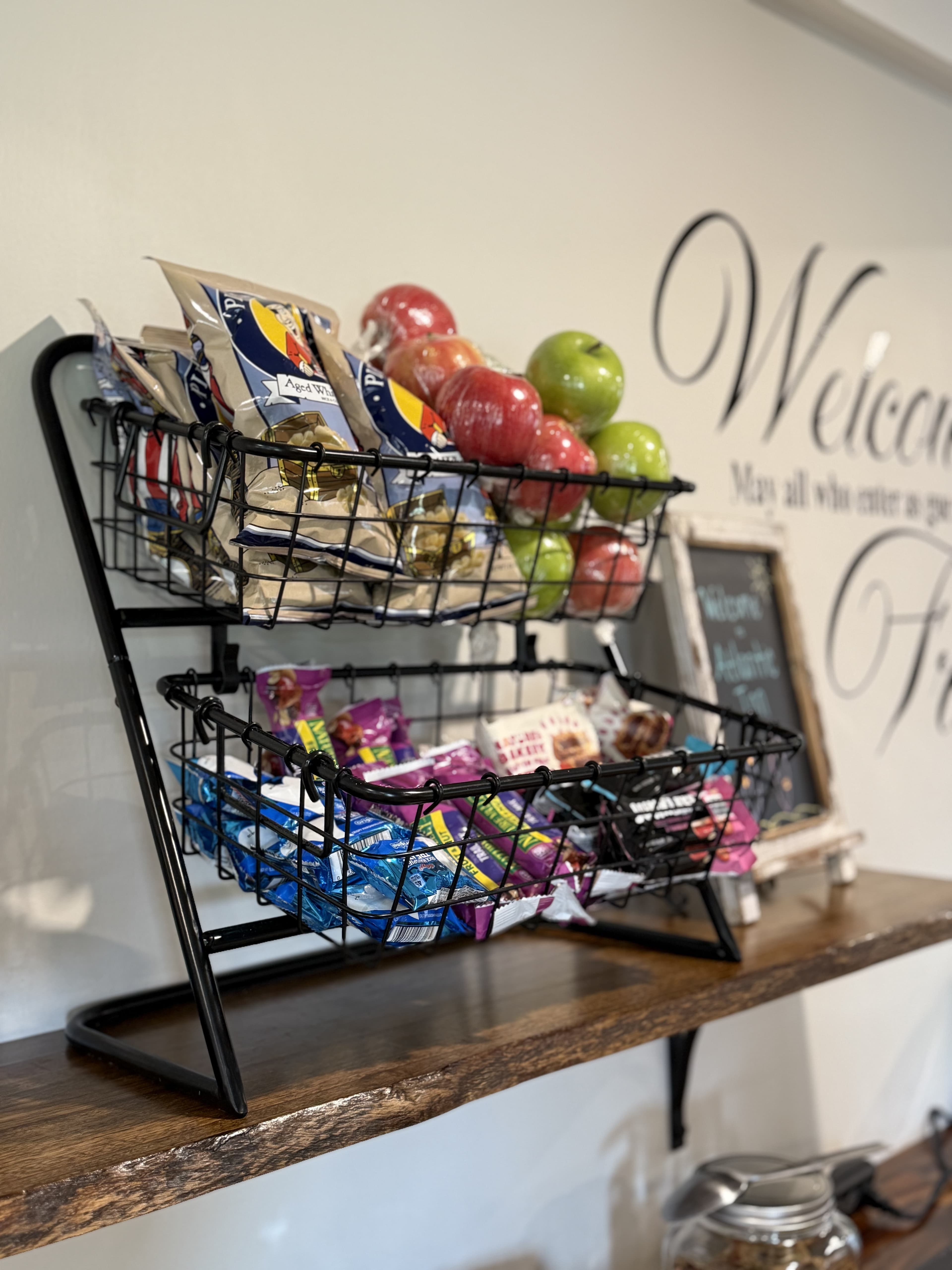 A wire basket display filled with snacks and apples on a wooden shelf against a welcoming wall.