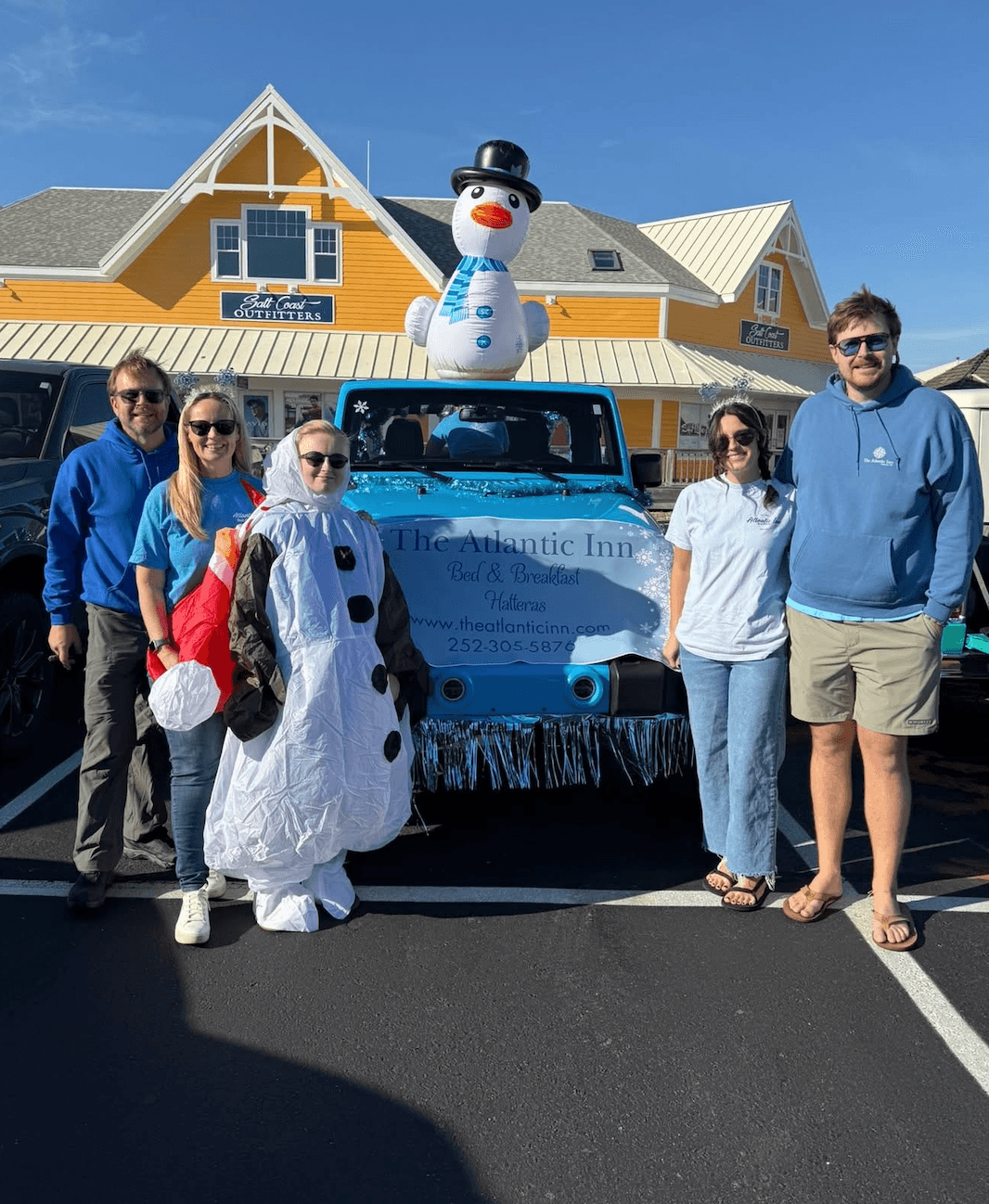 A group of five people poses in front of a festive blue Jeep decorated for the holidays, featuring a snowman on top.