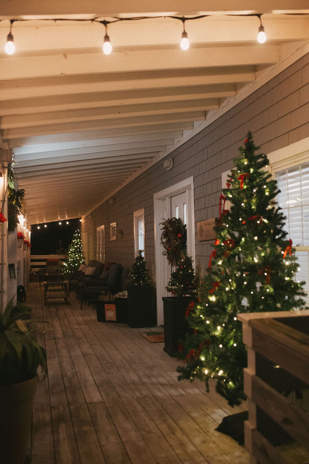 A warmly lit porch decorated for Christmas with trees and wreaths.
