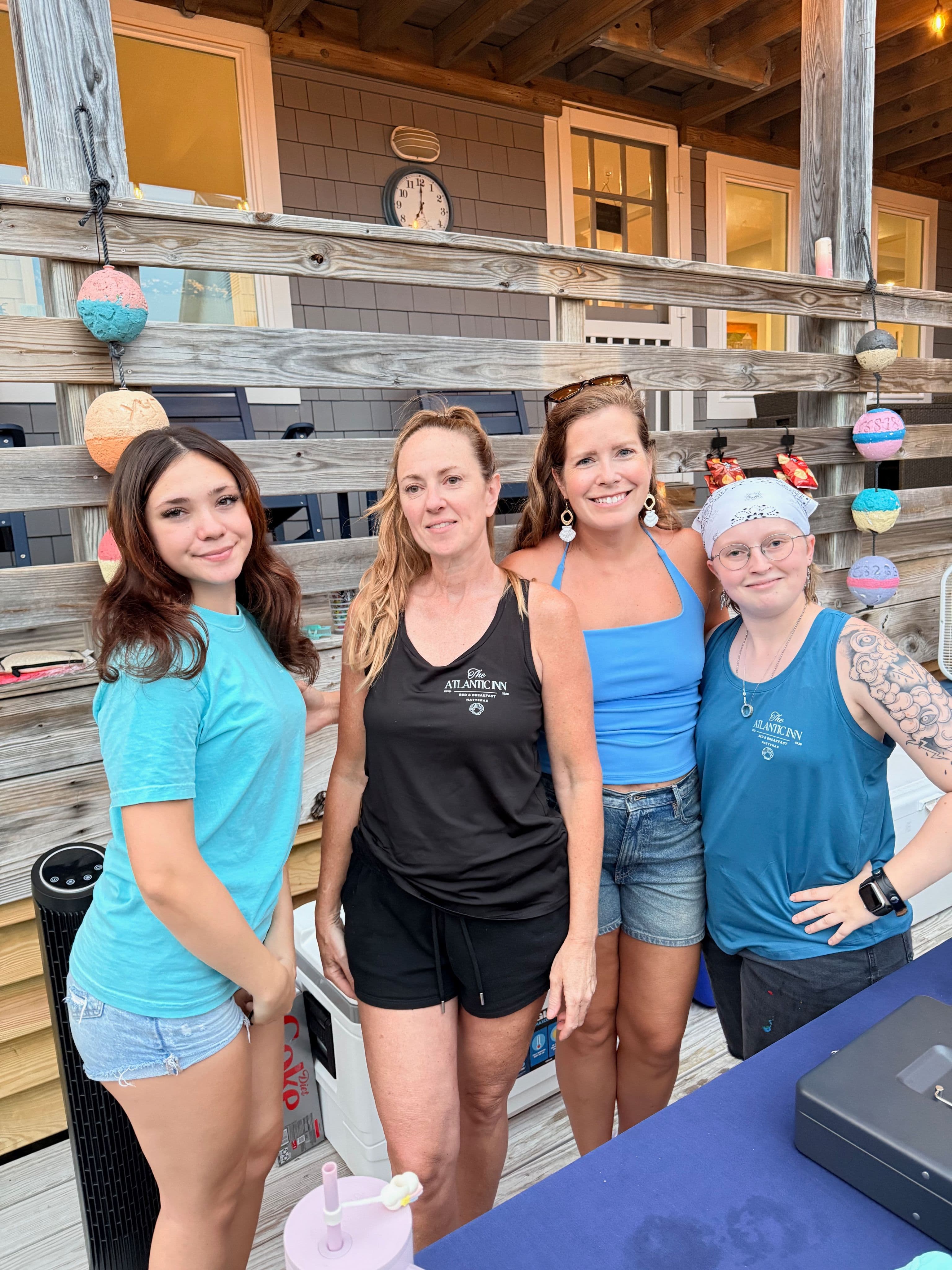 Four women stand together smiling in front of a wooden deck, with a clock and decorative lights in the background.