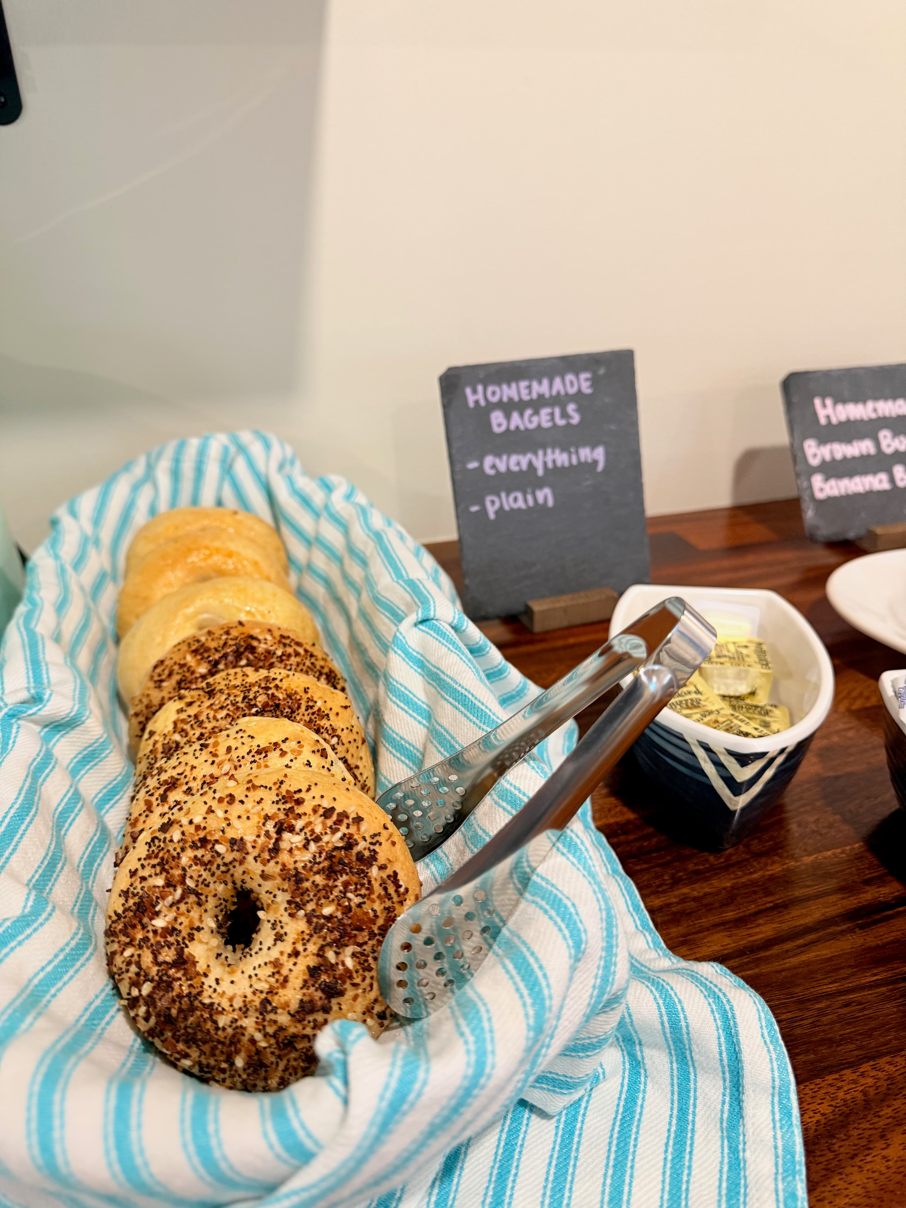 A basket of homemade bagels, featuring everything and plain varieties, with tongs for serving.