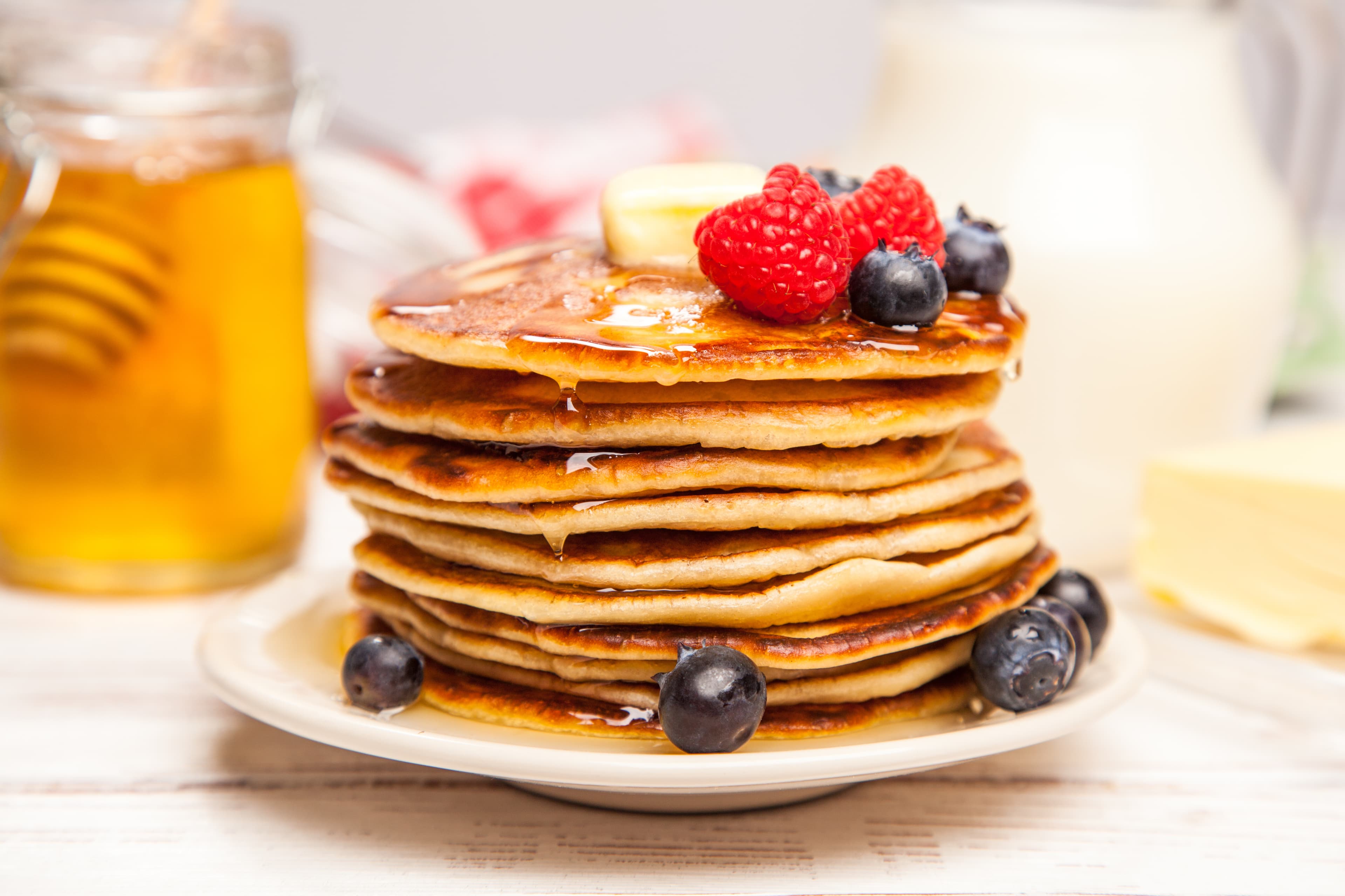 A stack of golden pancakes topped with berries and syrup on a plate, accompanied by honey and a glass of milk.