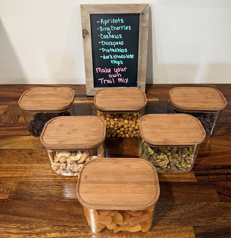 A selection of clear containers filled with various nuts and dried fruits, accompanied by a chalkboard sign inviting visitors to "Make your own Trail Mix."