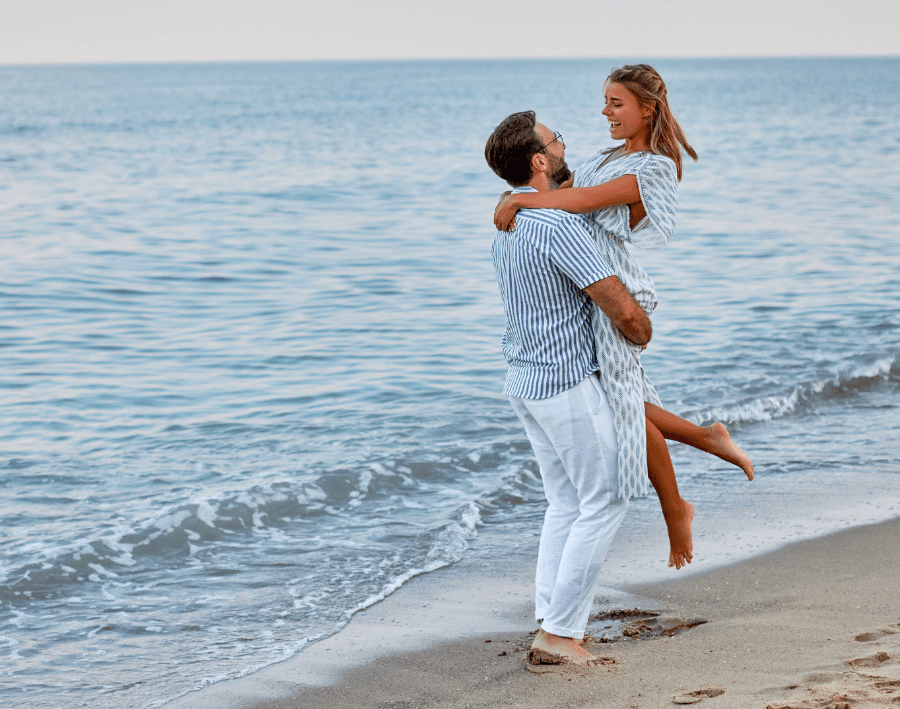 A man lifts a girl joyfully at the beach, with calm waves in the background.