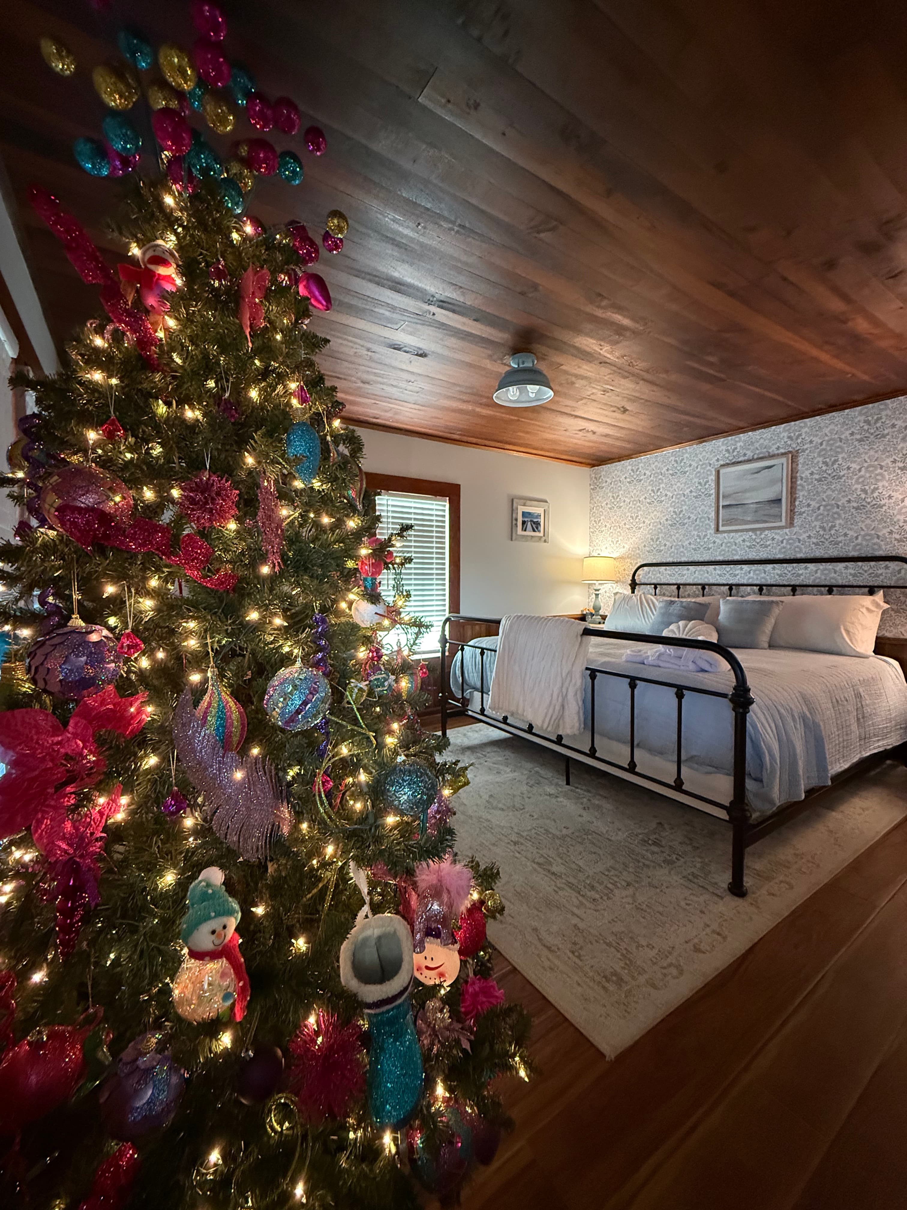 A brightly decorated Christmas tree stands in a cozy room featuring a bed and soft lighting.