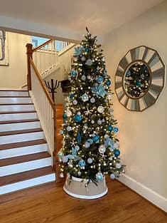 A beautifully decorated Christmas tree with blue and silver ornaments stands beside a staircase in a cozy entryway.