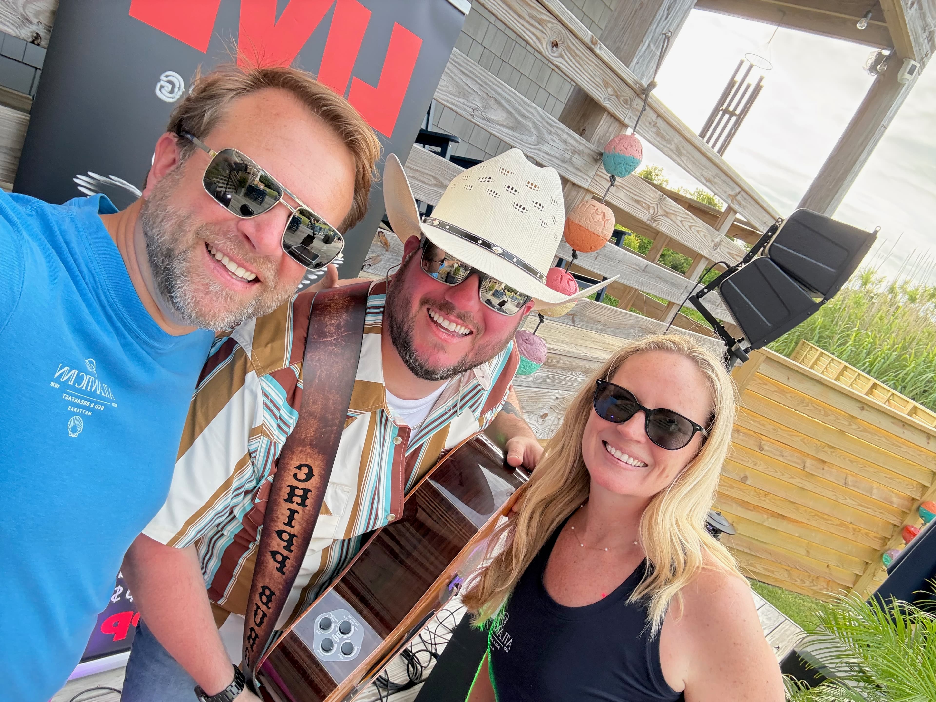 Three people, one wearing a cowboy hat, pose together with musical equipment in a casual outdoor setting.