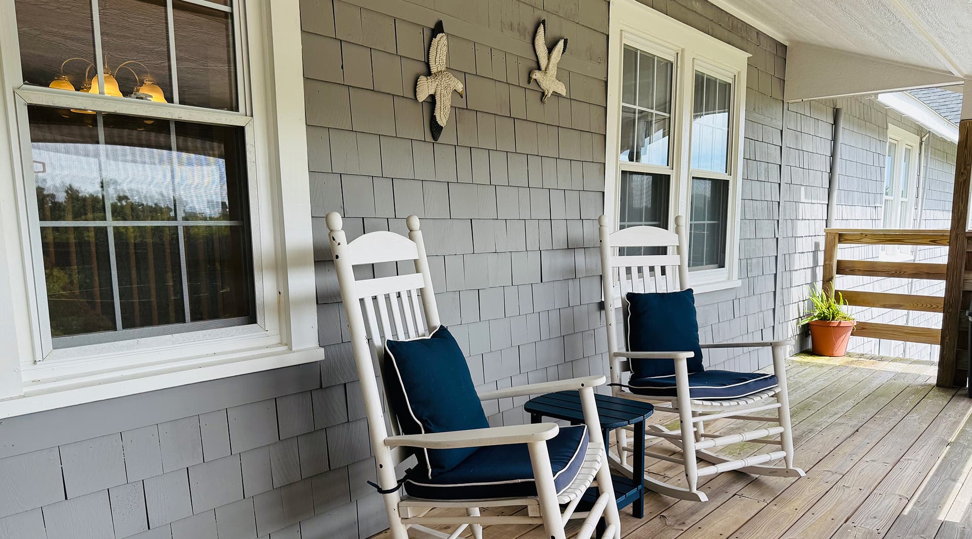 Two white rocking chairs with blue cushions on a wooden porch beside a gray shingled wall and windows.