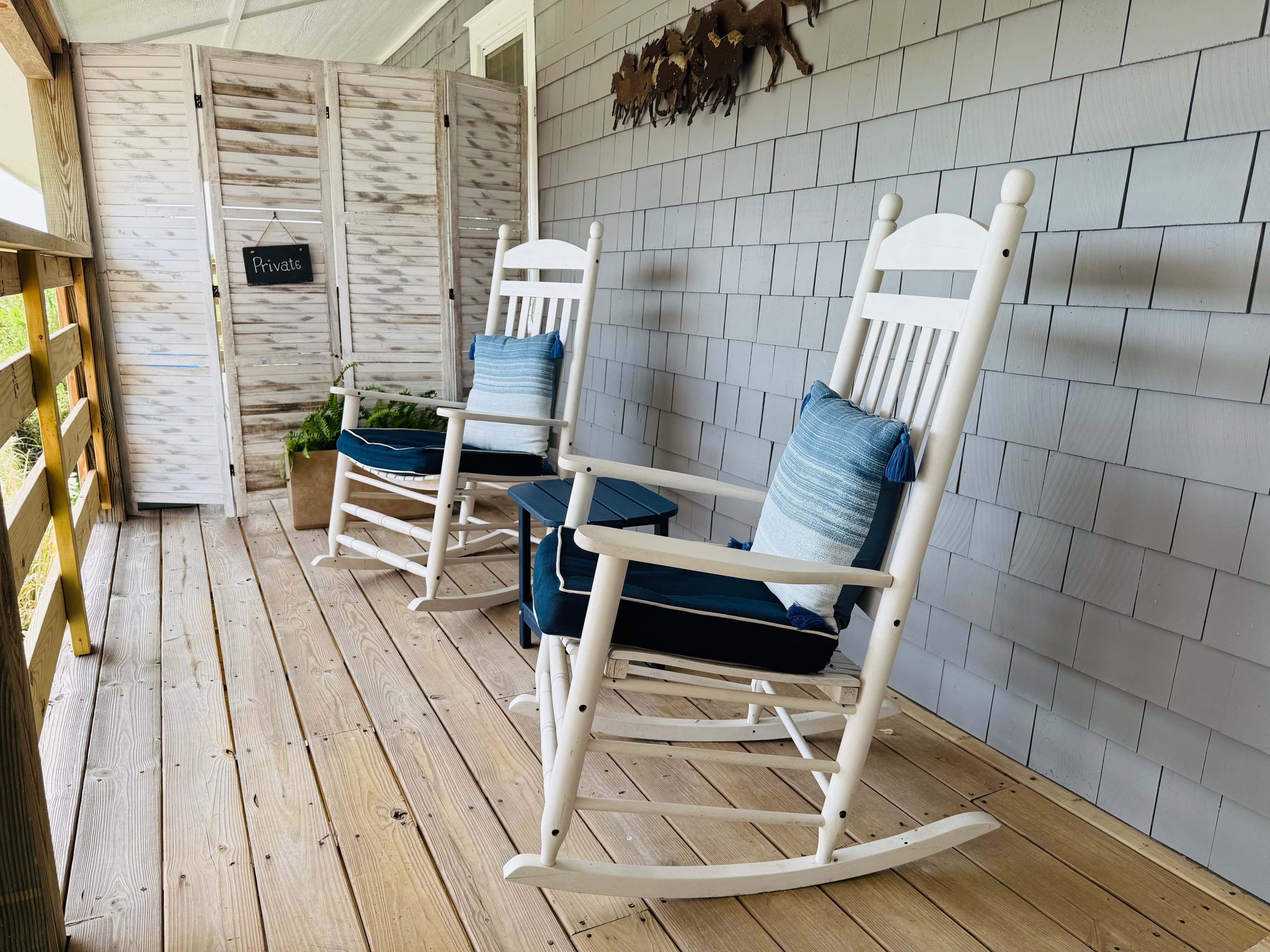 Two white rocking chairs with blue cushions on a wooden porch beside a privacy screen.