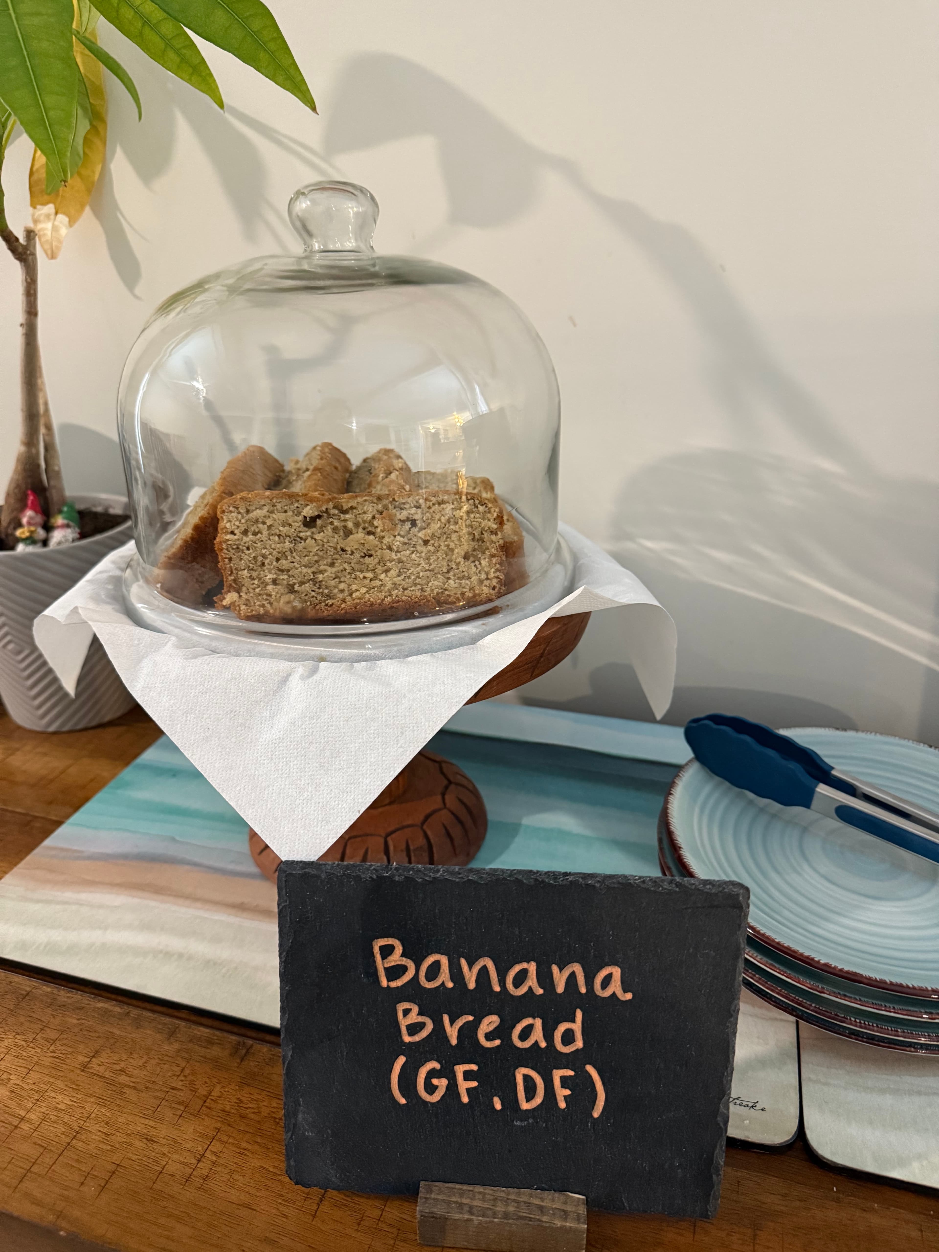 A glass dome covers slices of gluten-free, dairy-free banana bread on a wooden pedestal, with a sign identifying the item.