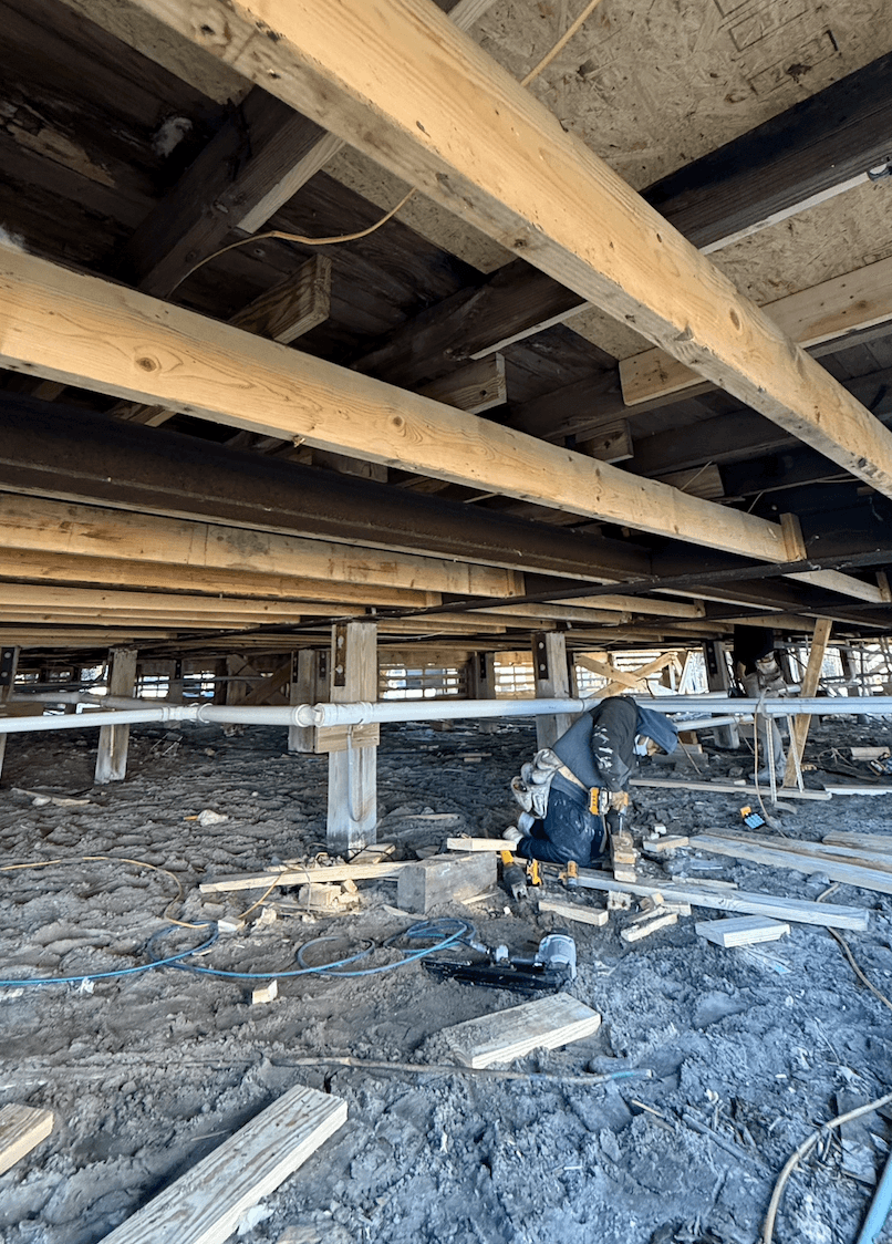 A construction worker is kneeling on the ground beneath a building, working with wooden beams and tools.
