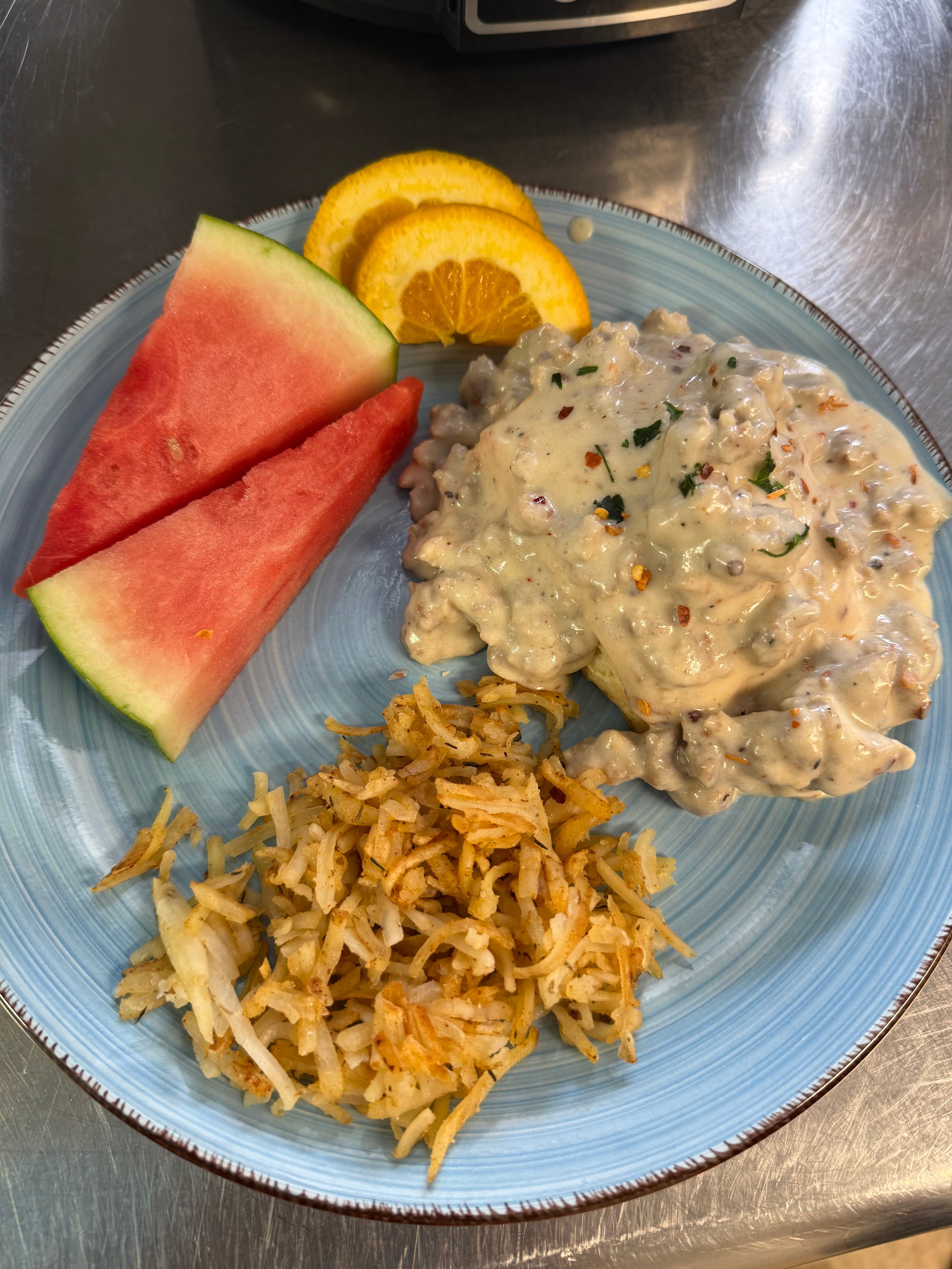 A blue plate featuring hashbrowns, creamy sausage gravy, watermelon, and orange slices.
