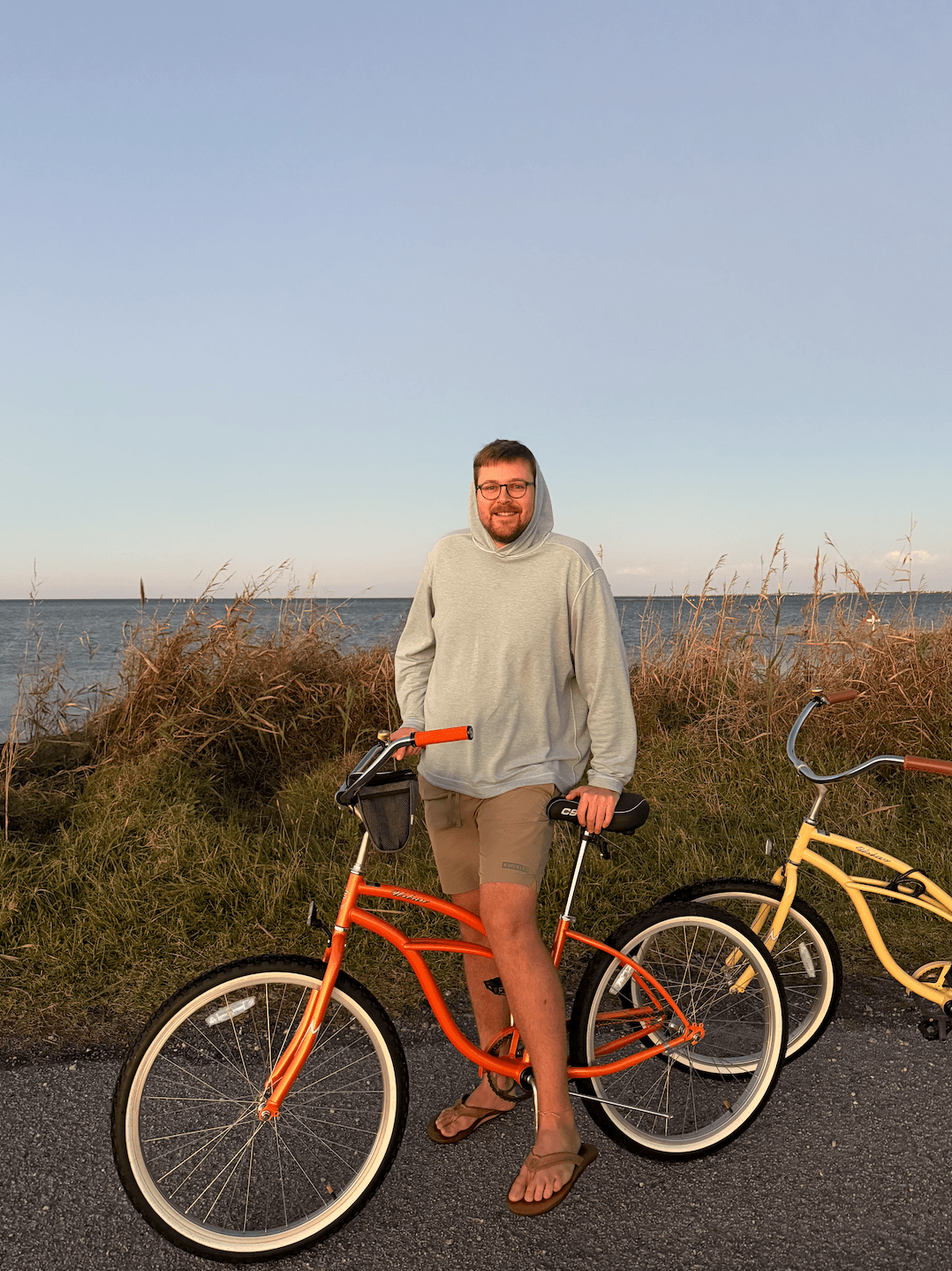 A man in a light green hoodie and shorts stands next to an orange bicycle by the water at sunset.