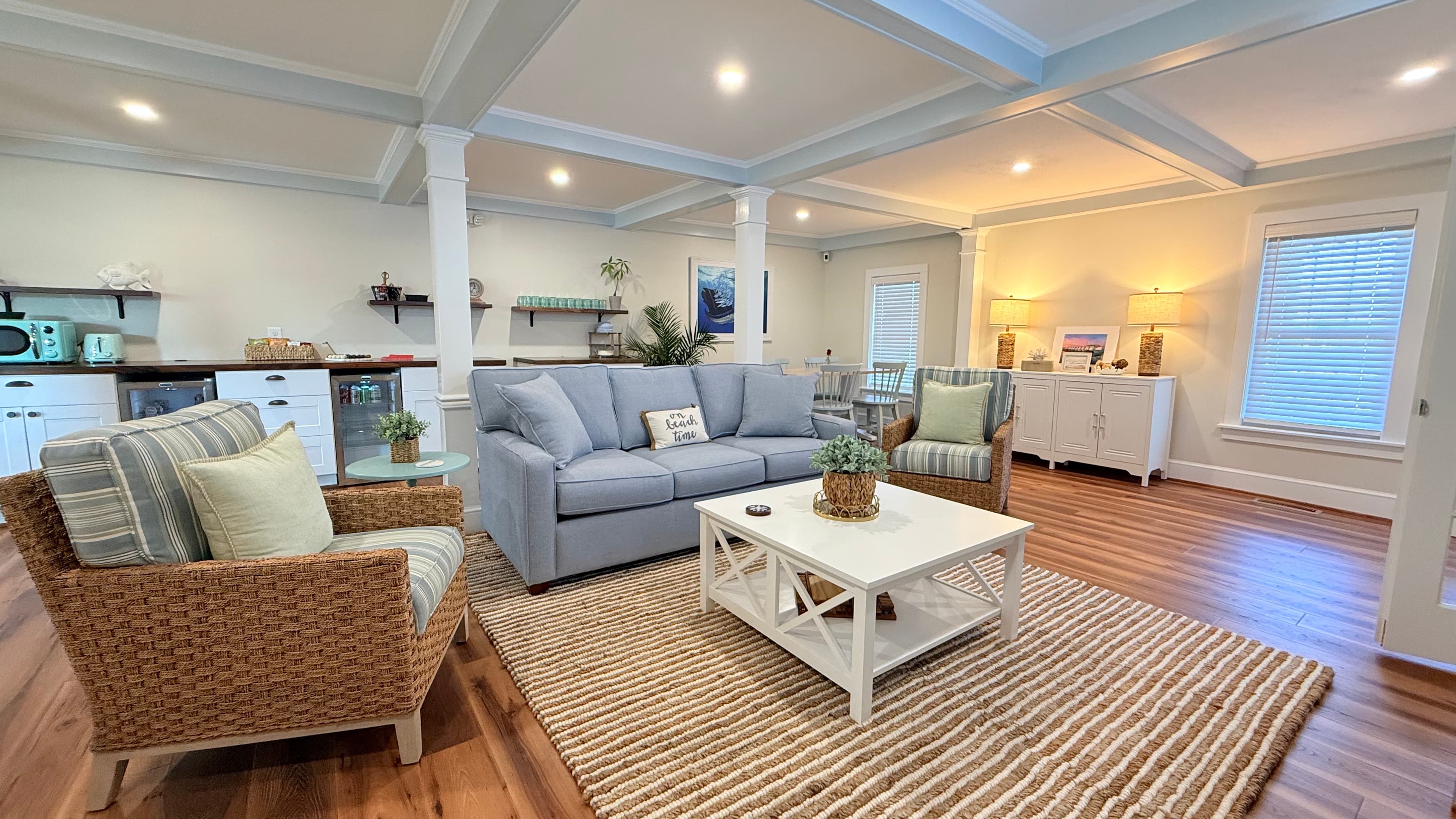 A cozy living room featuring a grey sofa, wicker chairs, a white coffee table, and natural light from nearby windows.