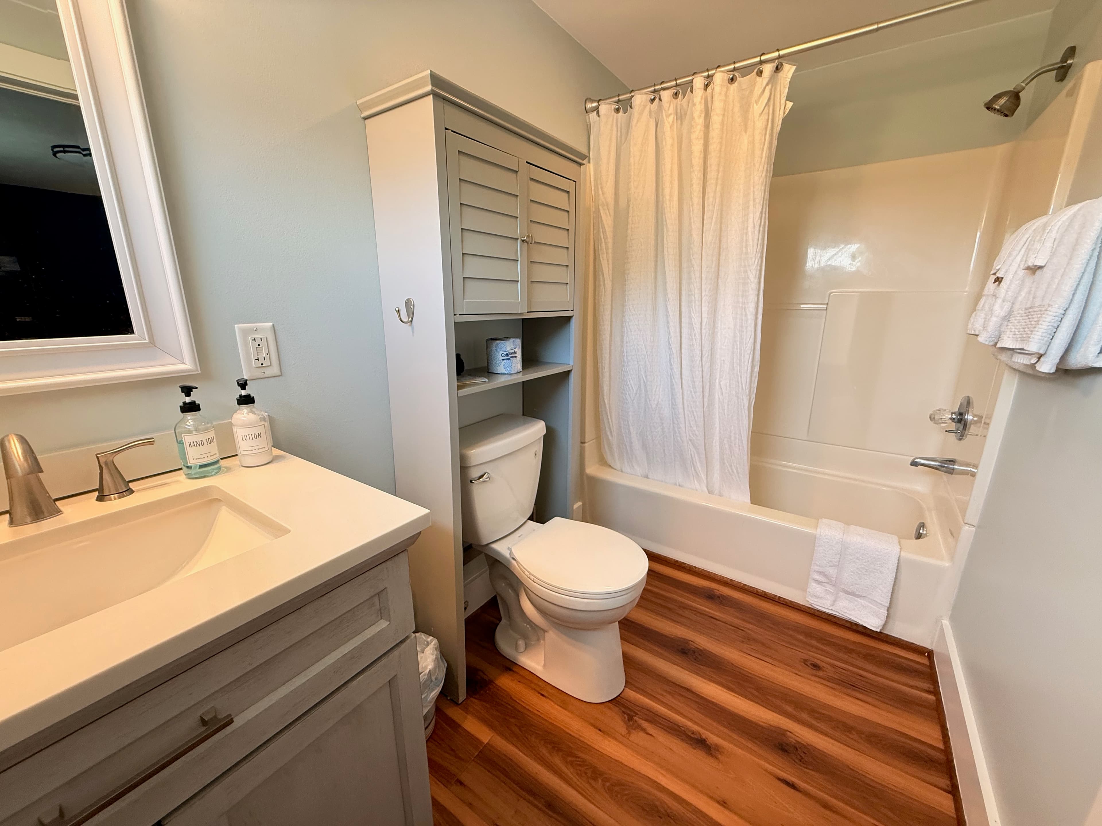 A brightly lit bathroom featuring a gray vanity with a white countertop and a white-framed mirror. The room has light gray walls, a small high window, and a modern light fixture with three clear glass globes.