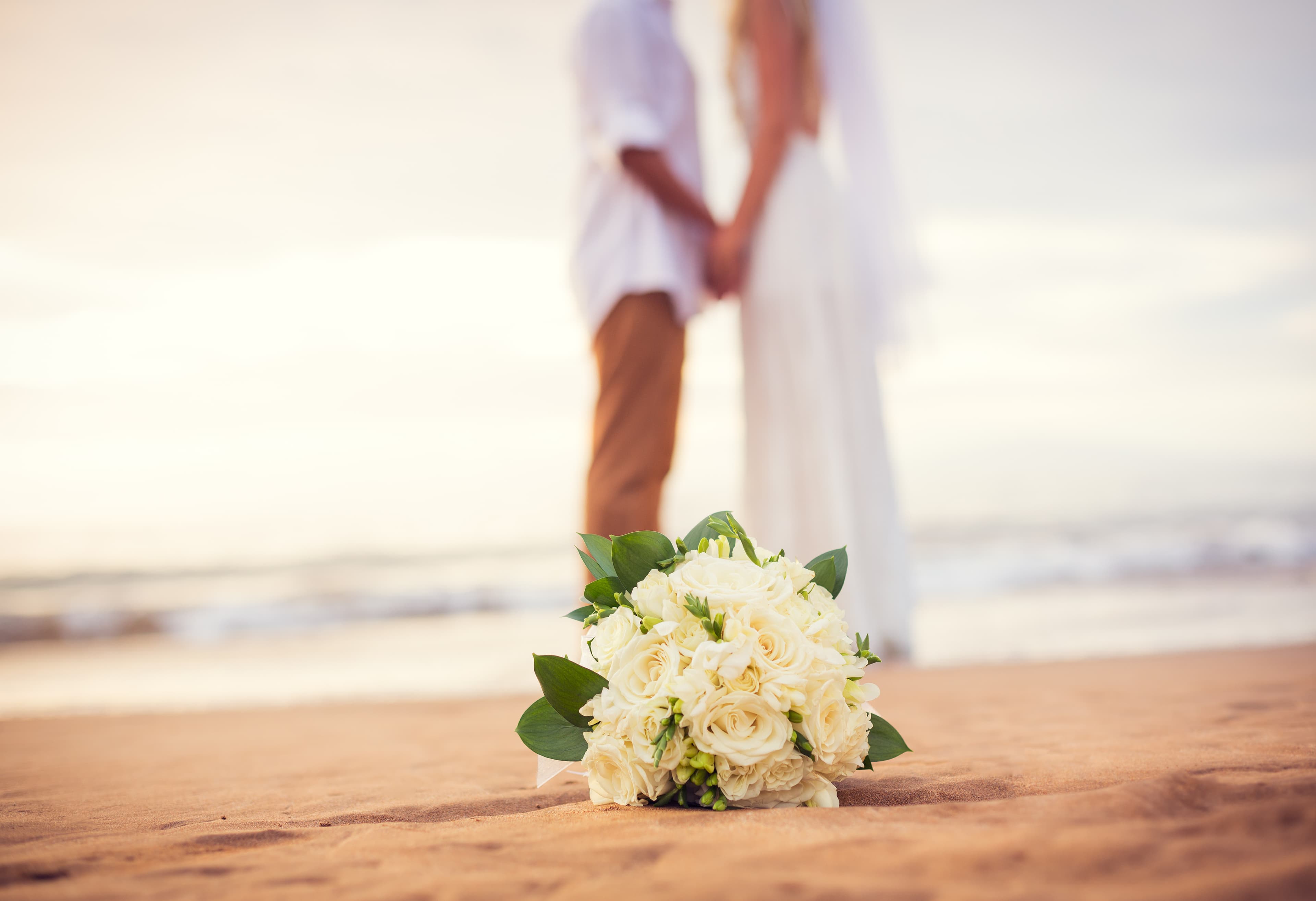 A bridal bouquet rests on the sand while a couple holds hands in the background.