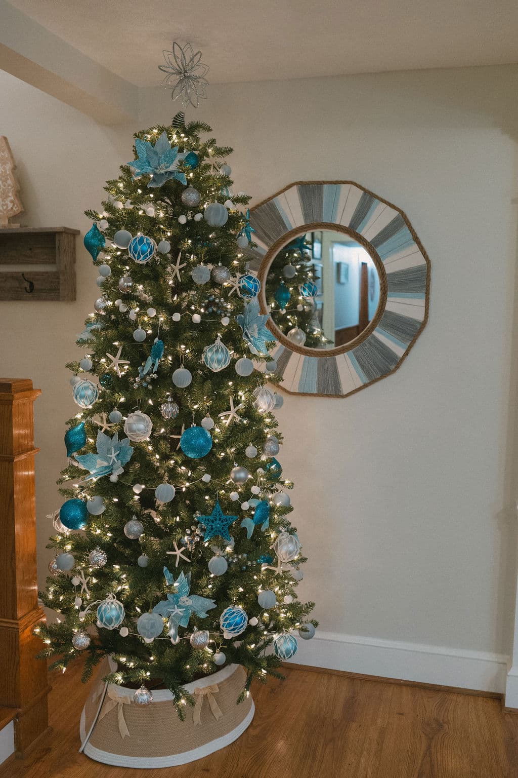 A decorated Christmas tree with blue and silver ornaments stands beside a round mirror in a cozy room.