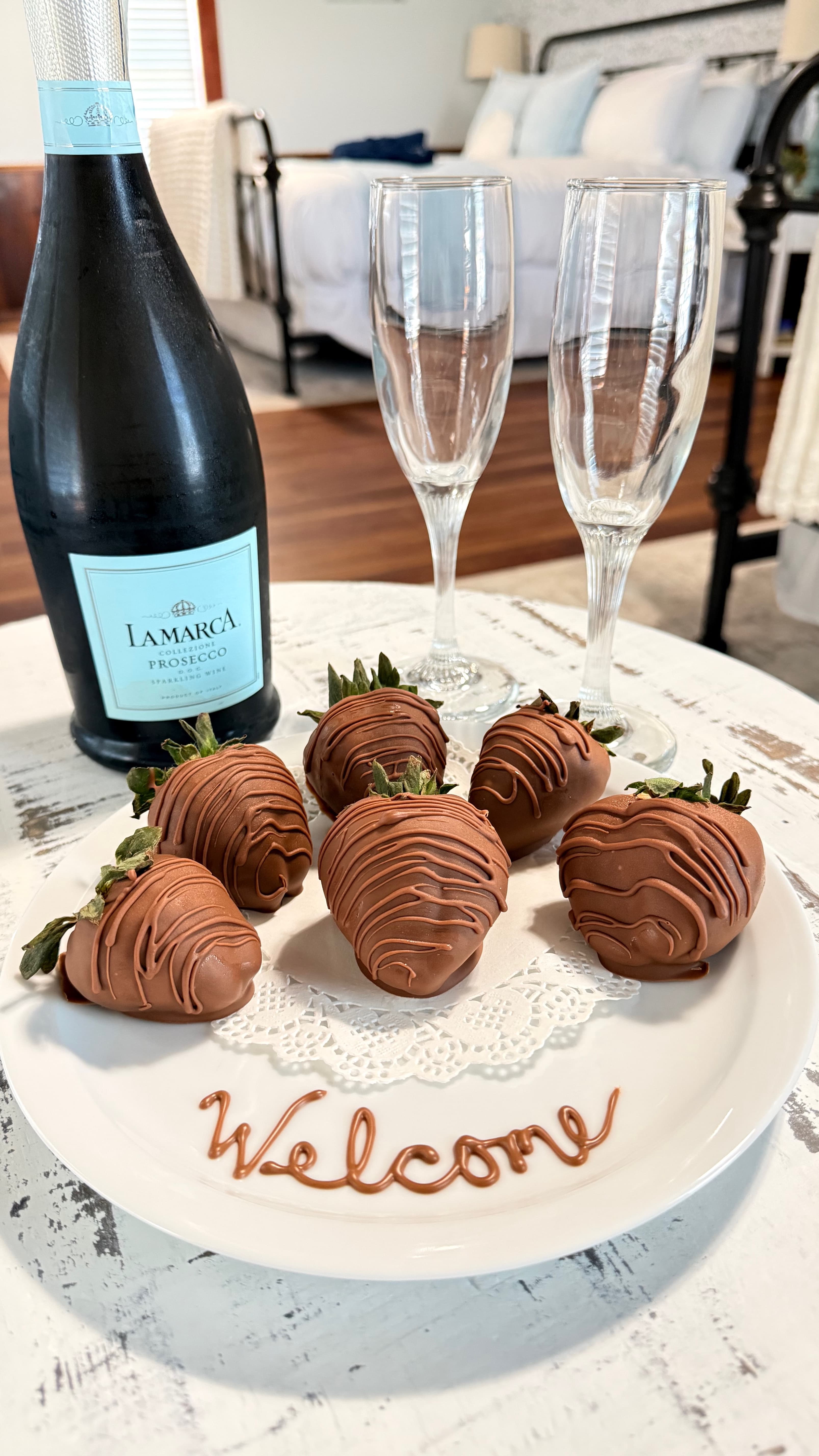A plate of chocolate-covered strawberries with a bottle of Prosecco and two champagne flutes, decorated with "Welcome" written in chocolate.