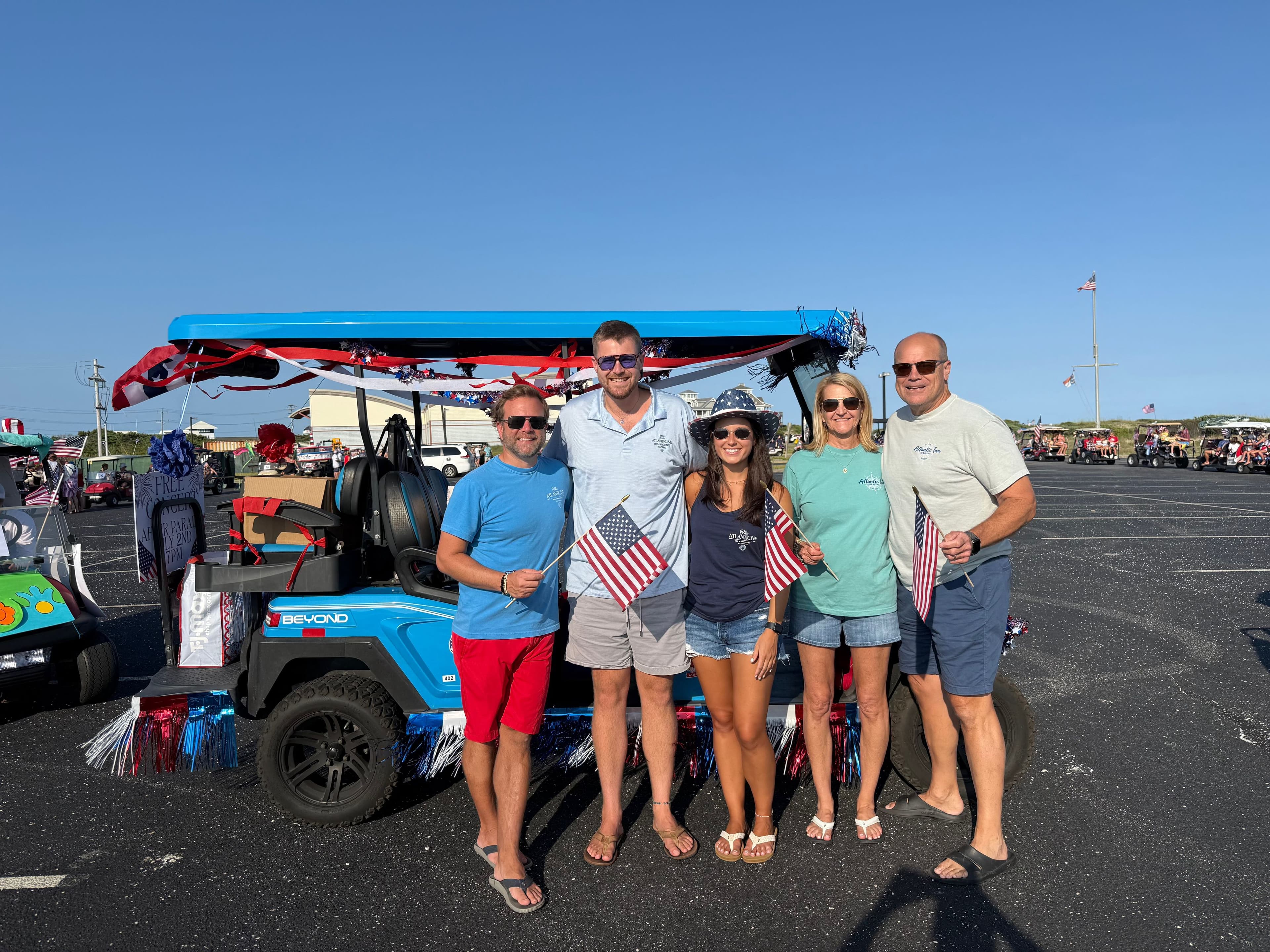 A group of five people holding small American flags stands in front of a decorated golf cart on a sunny day.